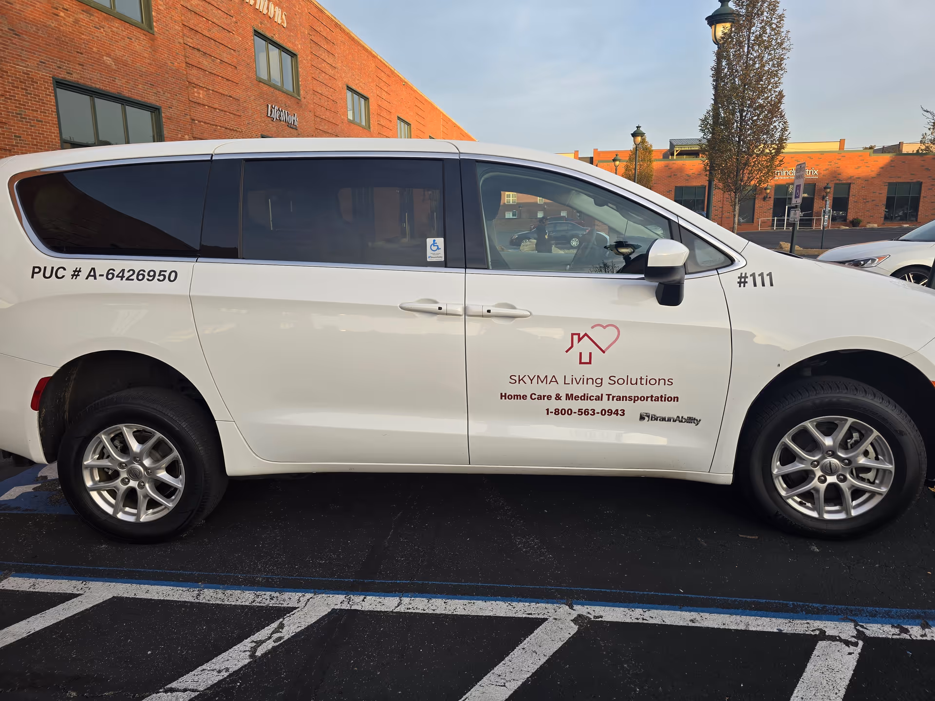 A white SKYMA Living Solutions van parked in a lot beside a brick building exterior.