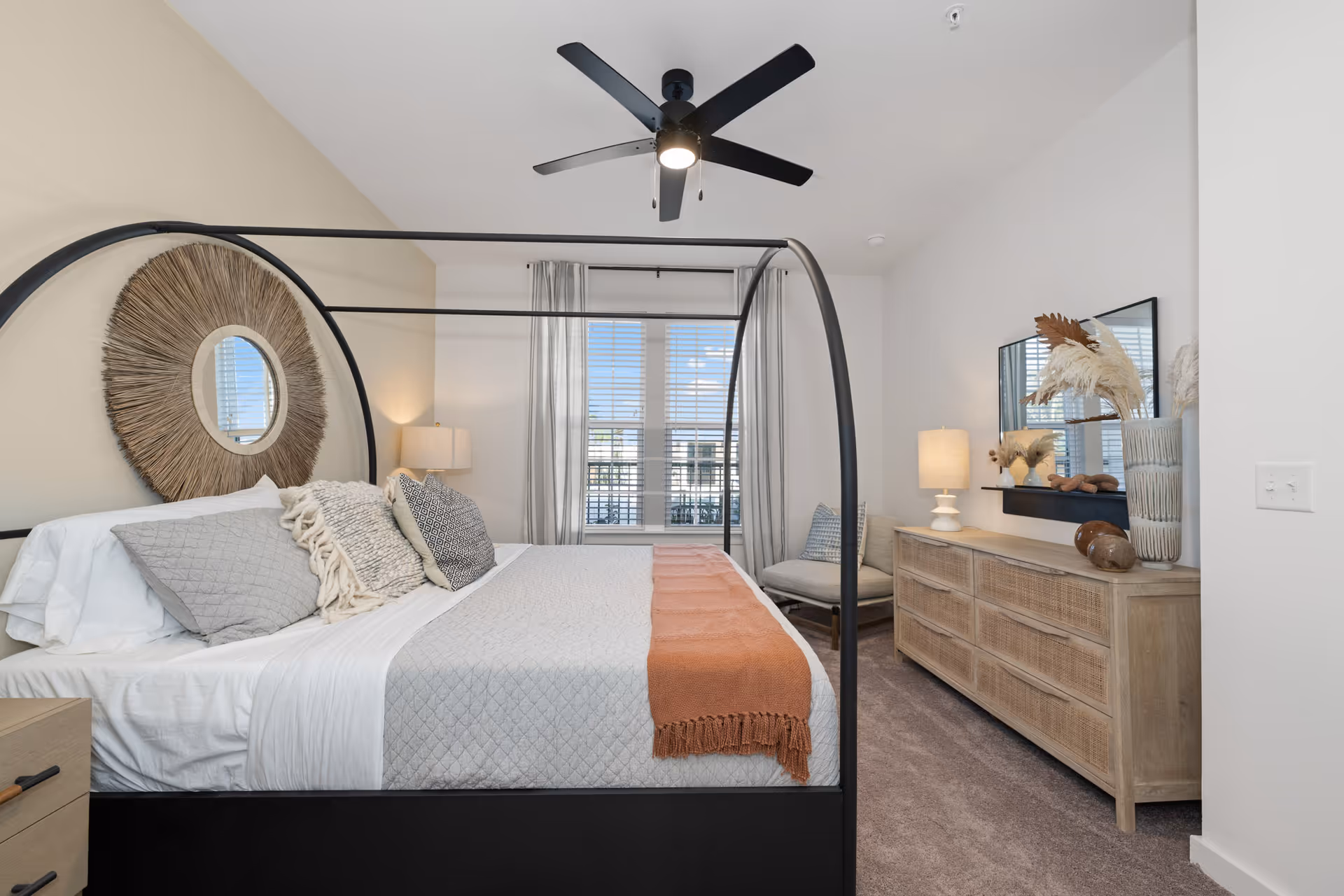A modern bedroom with a black metal canopy bed adorned with gray and patterned pillows and a rust-colored throw blanket. The room features a large round decorative mirror above the bed, a ceiling fan with light, a window with sheer curtains, a wooden dresser with a lamp and decorative items, and a cushioned chair in the corner.