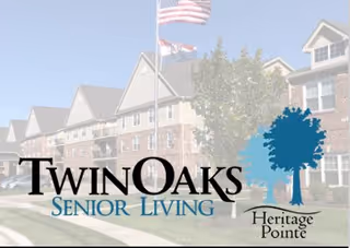 Exterior view of a senior living facility building with multiple peaked roofs, brick and siding facade, and an American flag flying on a flagpole in front. The image includes the Twin Oaks Senior Living and Heritage Pointe logos overlaid.