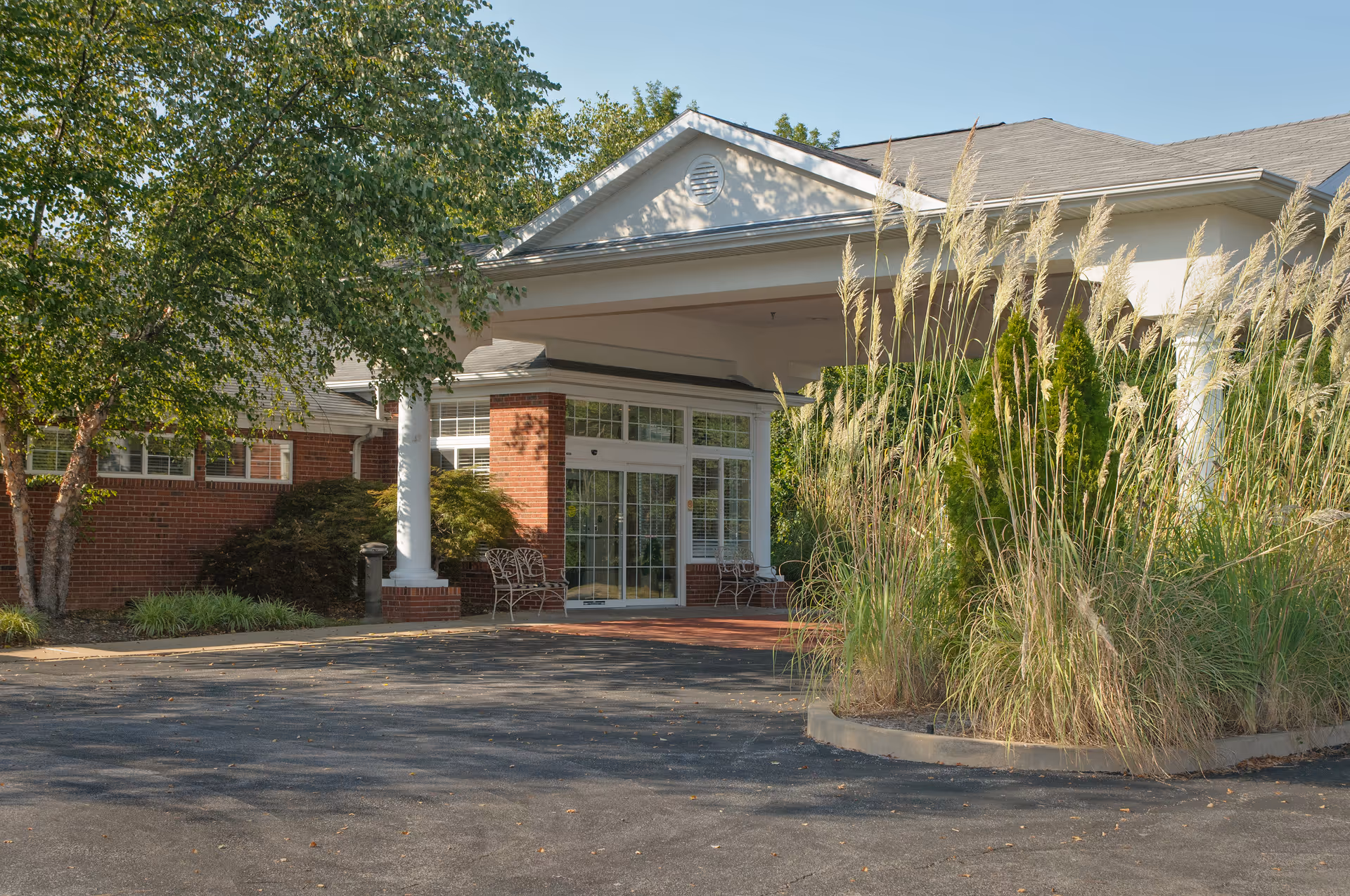 Covered front entrance of a brick senior living building with white columns, glass doors, benches, and surrounding trees and ornamental grasses.