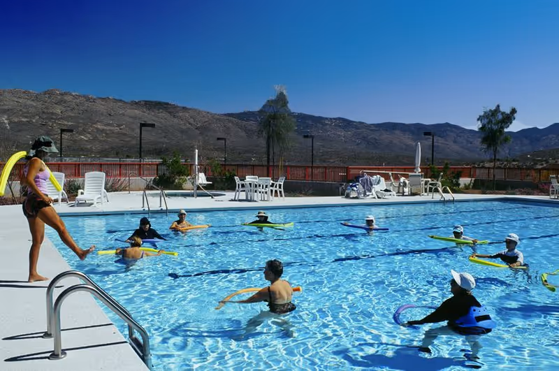 A group of people participating in a water exercise class in an outdoor swimming pool with mountains in the background under a clear blue sky. An instructor stands at the poolside demonstrating movements.