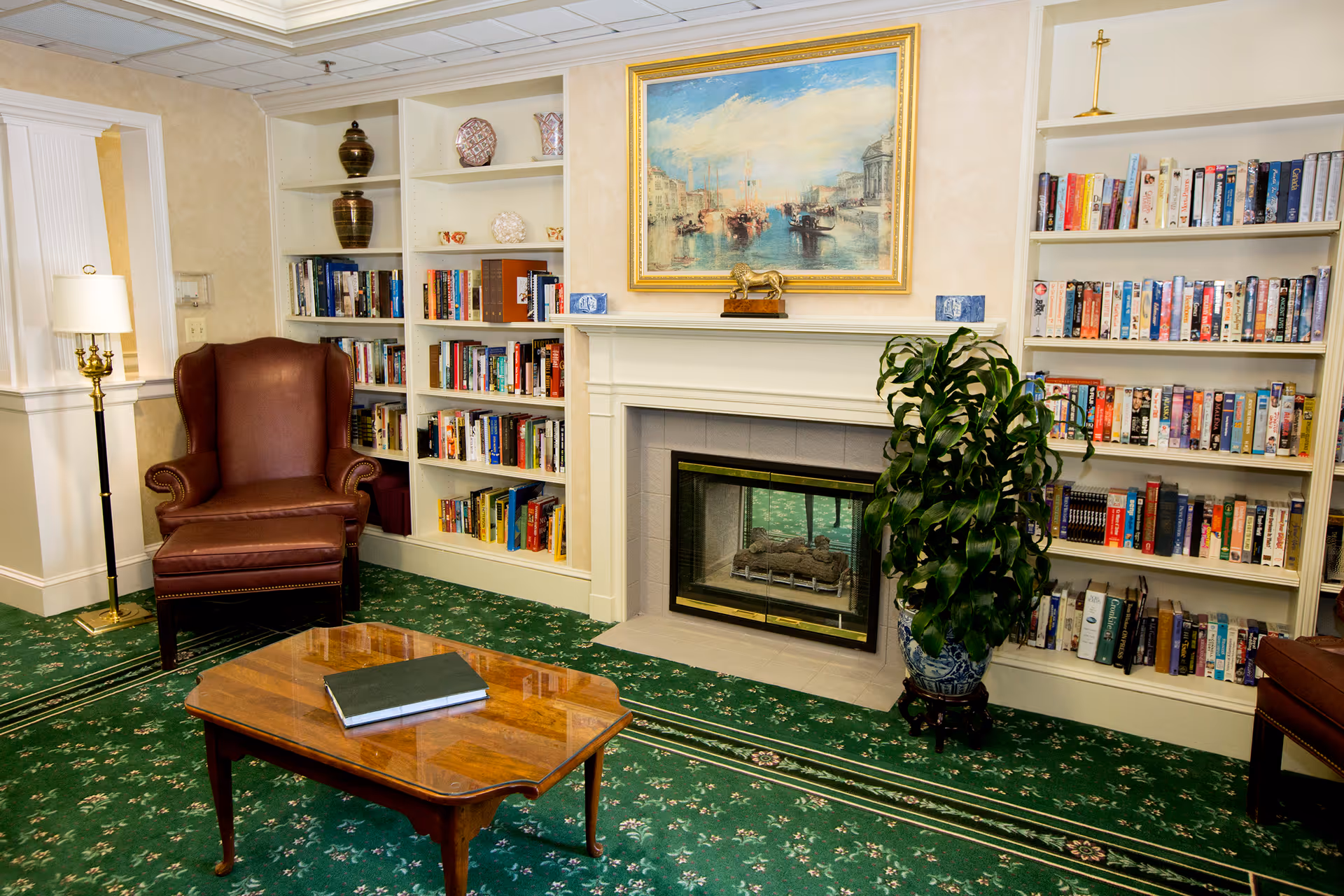 A cozy living room area with a green floral carpet, a brown leather armchair, a wooden coffee table with a closed book on it, built-in white bookshelves filled with books and decorative items, a fireplace with a glass front, a potted plant in a decorative pot, a floor lamp with a white shade, and a framed painting of a harbor scene above the fireplace.