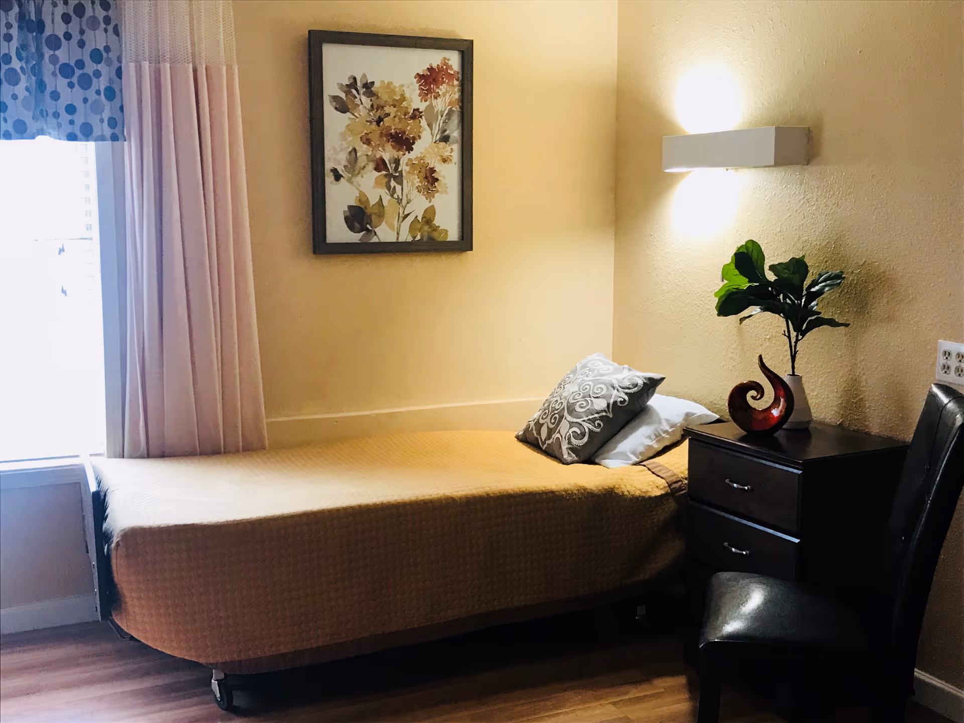 A small, cozy bedroom with a single bed covered in a yellow blanket and two pillows. Next to the bed is a dark wooden nightstand with two drawers, a decorative red sculpture, and a potted plant. A black chair is placed beside the nightstand. The room has light yellow walls, a framed floral painting above the bed, and a window with pink curtains and a blue valance with polka dots.