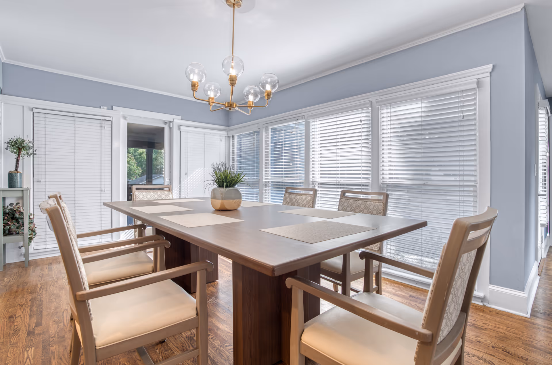 Bright dining room with a long wooden table, eight cushioned chairs, and large windows.