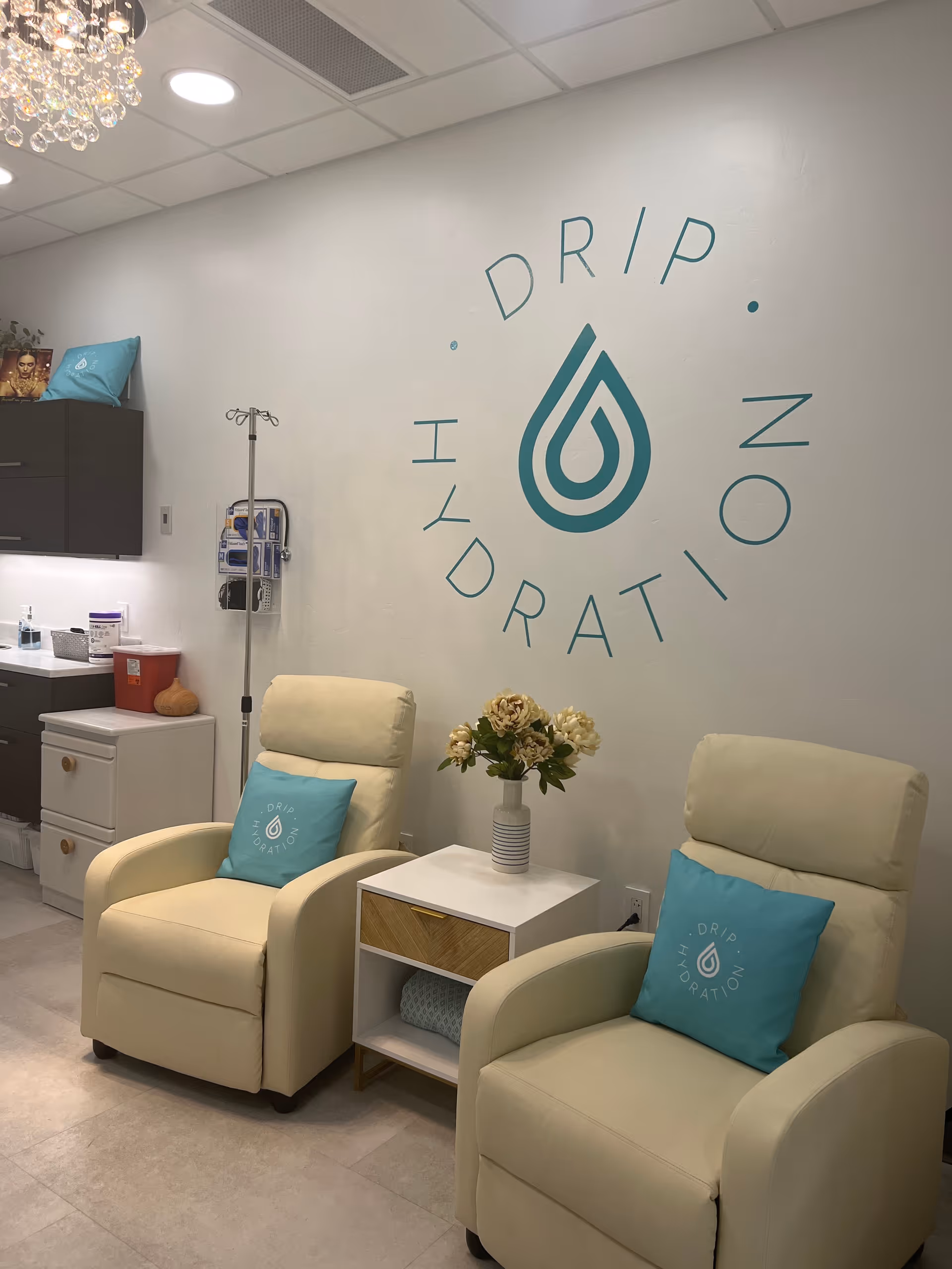 Clinic waiting area with two beige chairs, IV stand, side table, and Drip Hydration logo.