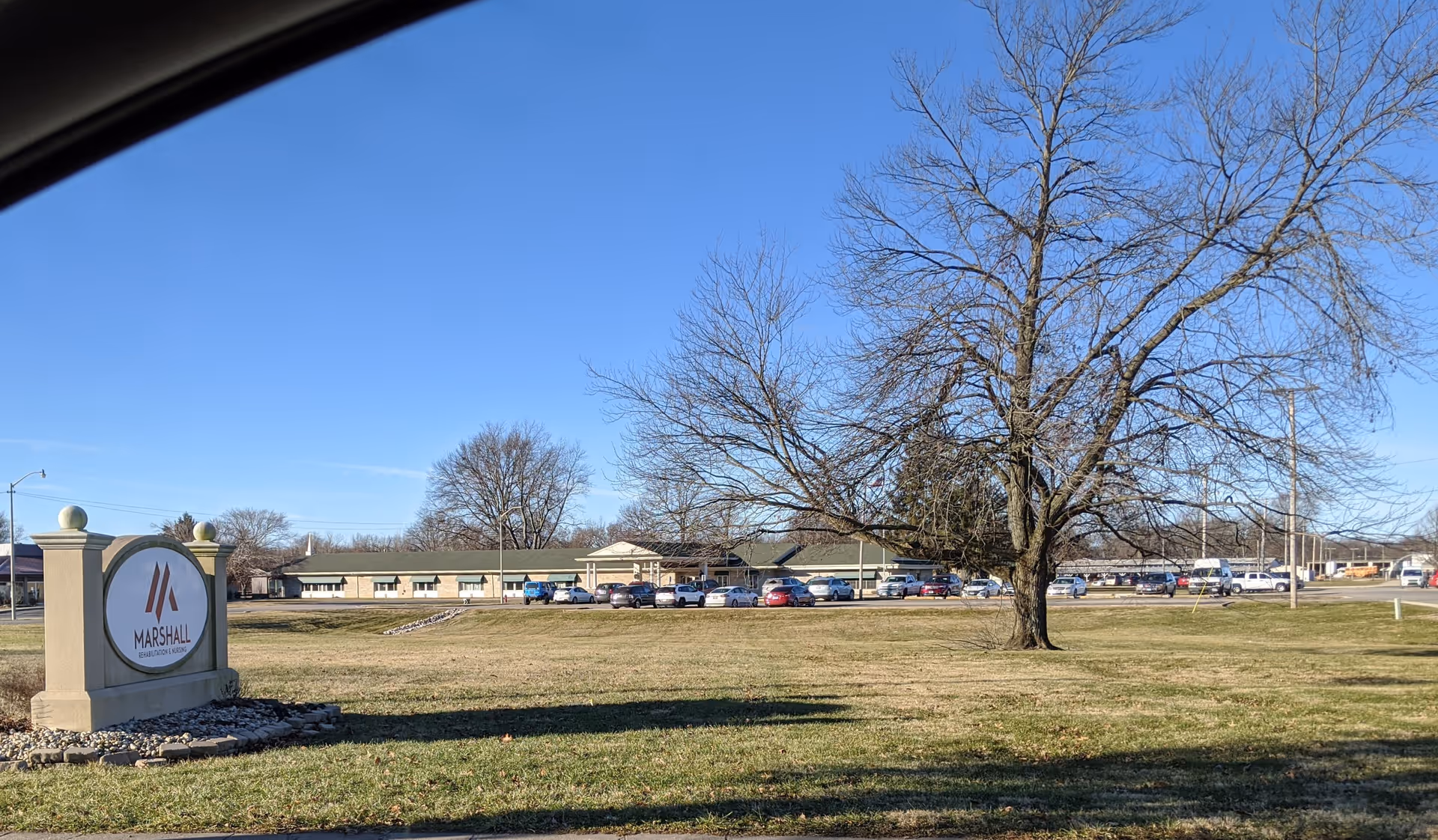 Exterior view of Marshall Rehabilitation and Nursing facility with a large sign in the foreground on a grassy area, a leafless tree, and a parking lot with several cars in front of a long, single-story building under a clear blue sky.