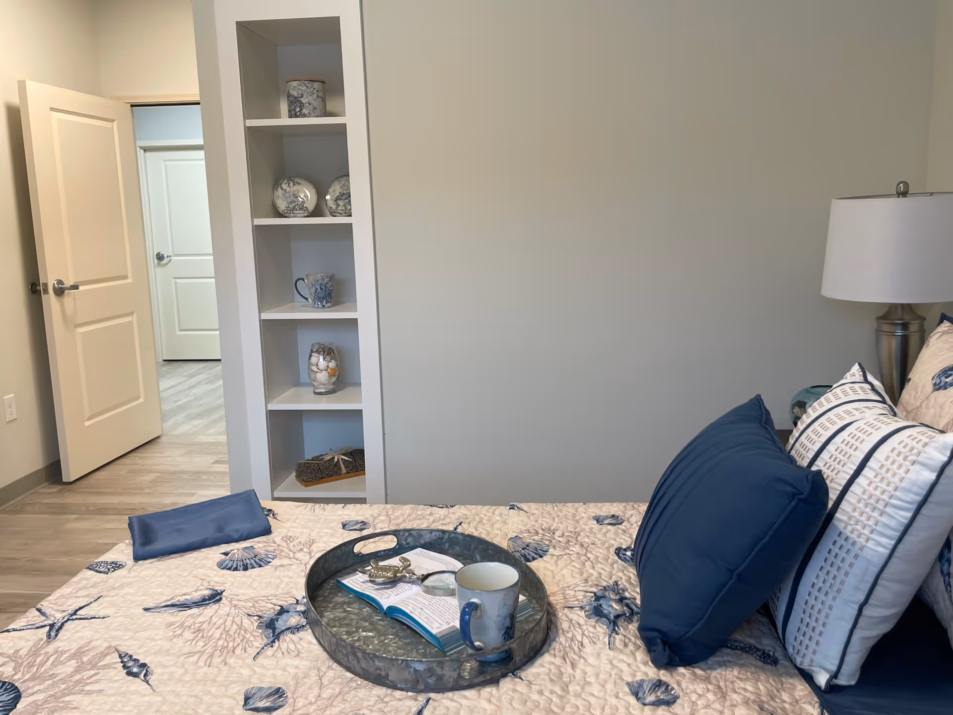 Blue-themed bedroom with seashell quilt, pillows, tray, mug, and built-in shelving.