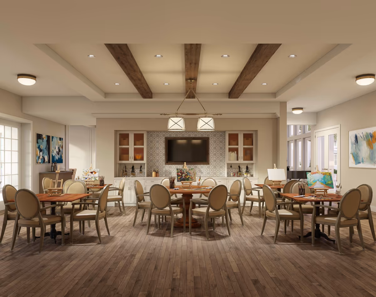 Bright communal dining and activity room with round wooden tables and beige upholstered chairs, cabinetry and a wall-mounted TV under decorative ceiling beams.