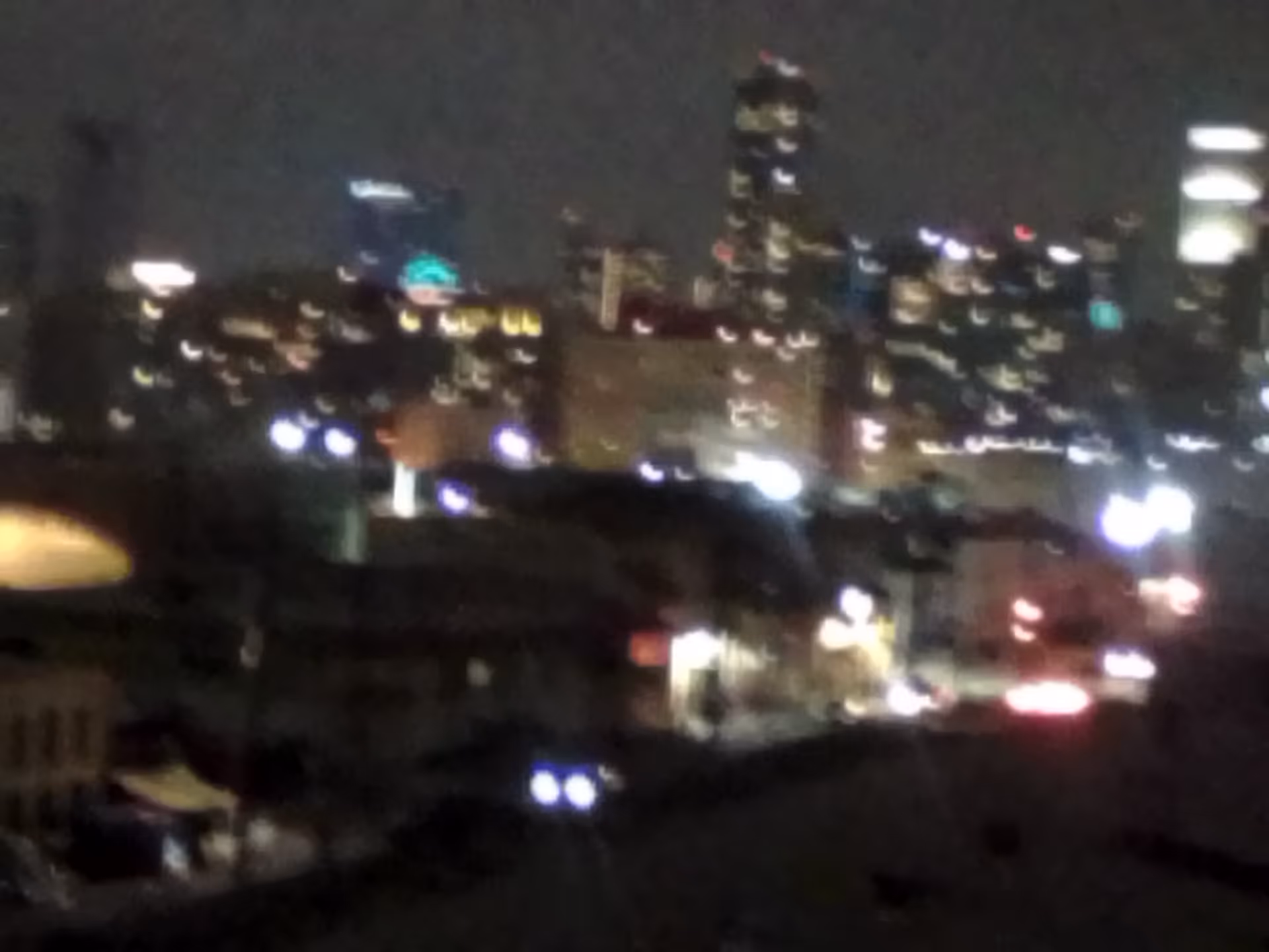 Blurry nighttime cityscape with bright building lights and street lamps.