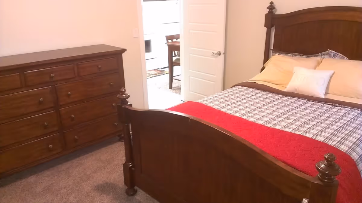 Wood-framed bed with plaid bedding and a red throw in a bedroom with a matching wooden dresser and an open door to another room.