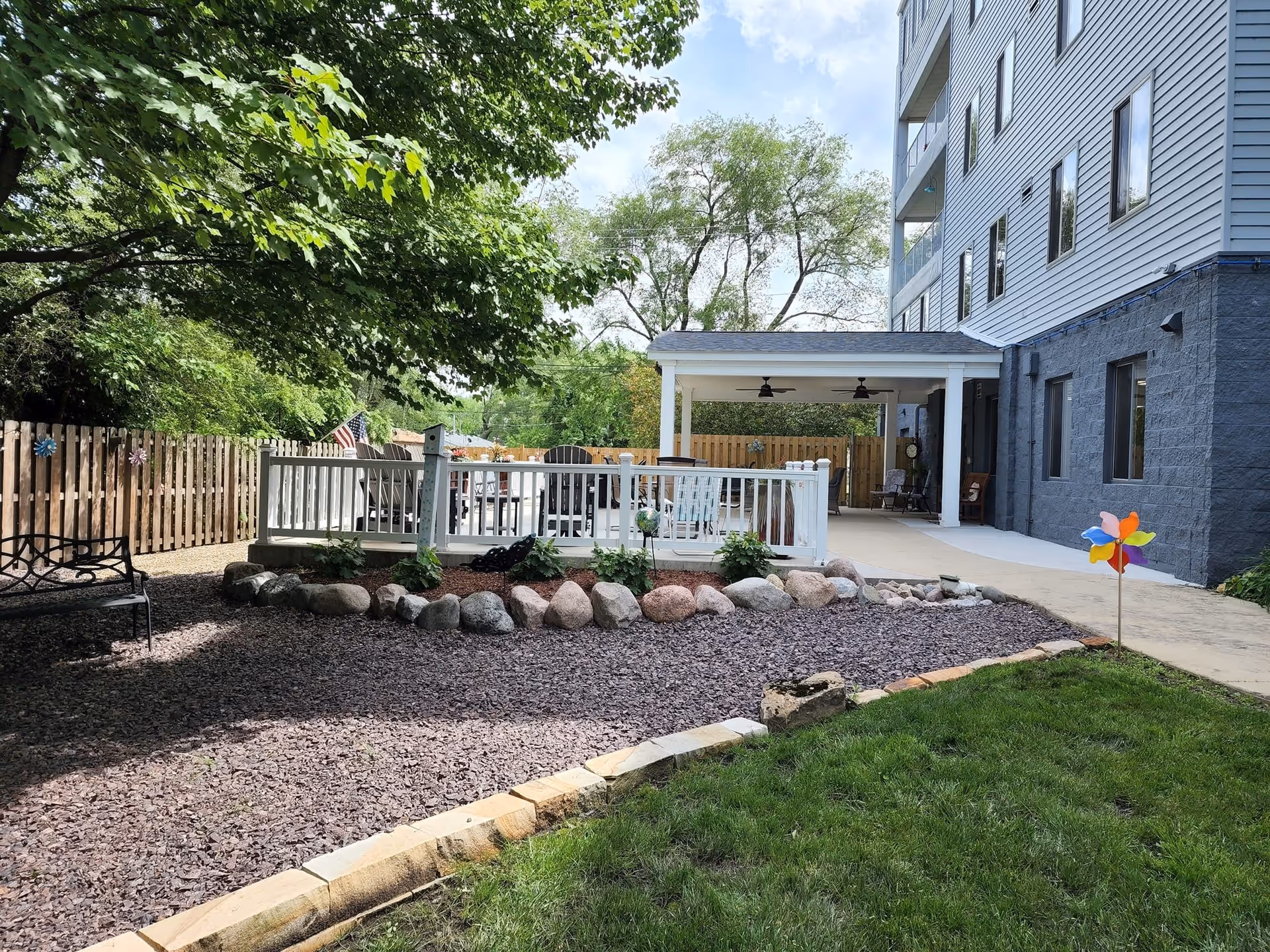 Outdoor garden area at Springfield Supportive Living Center with a gravel path, green grass, a wooden fence, and a covered patio attached to a multi-story building. There is a black metal bench under a tree on the left and a colorful pinwheel on the right side of the image.