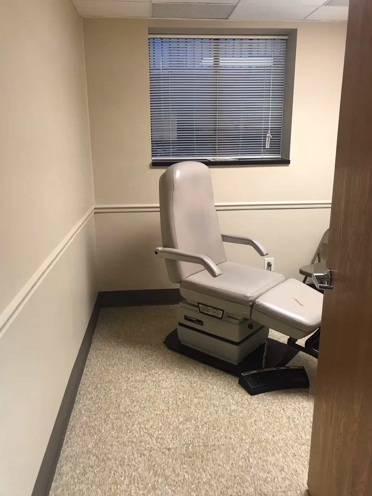 Medical examination chair in a small beige-walled room with a window and blinds.