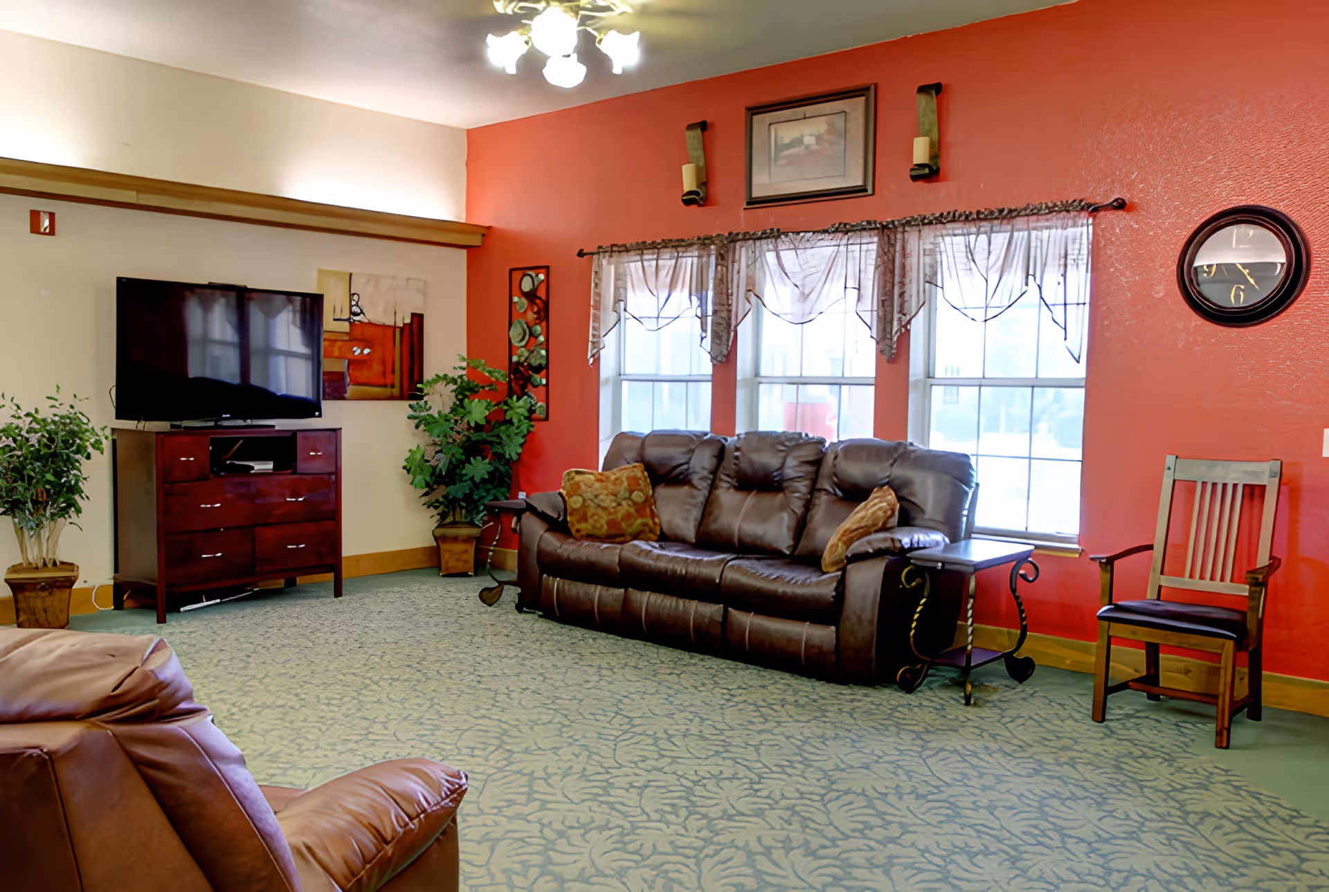 A cozy living room with a dark brown leather sofa and matching armchair, a wooden TV stand with a flat-screen television, two potted plants, a wall clock, and three large windows with sheer curtains. The walls are painted red and beige, and the floor is covered with a patterned green carpet.