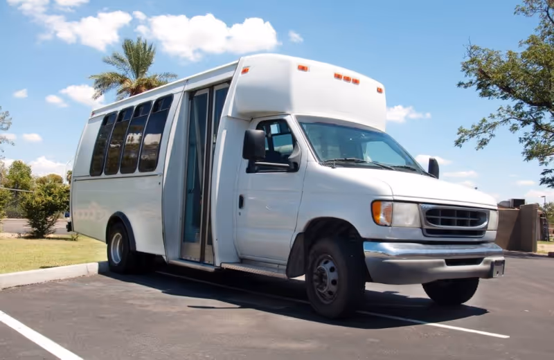 White shuttle bus with open door parked in a lot beside trees.