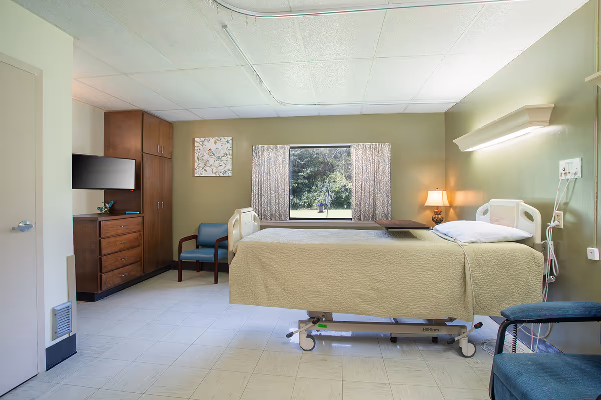 Hospital-style patient room with bed, dresser, window, and seating.