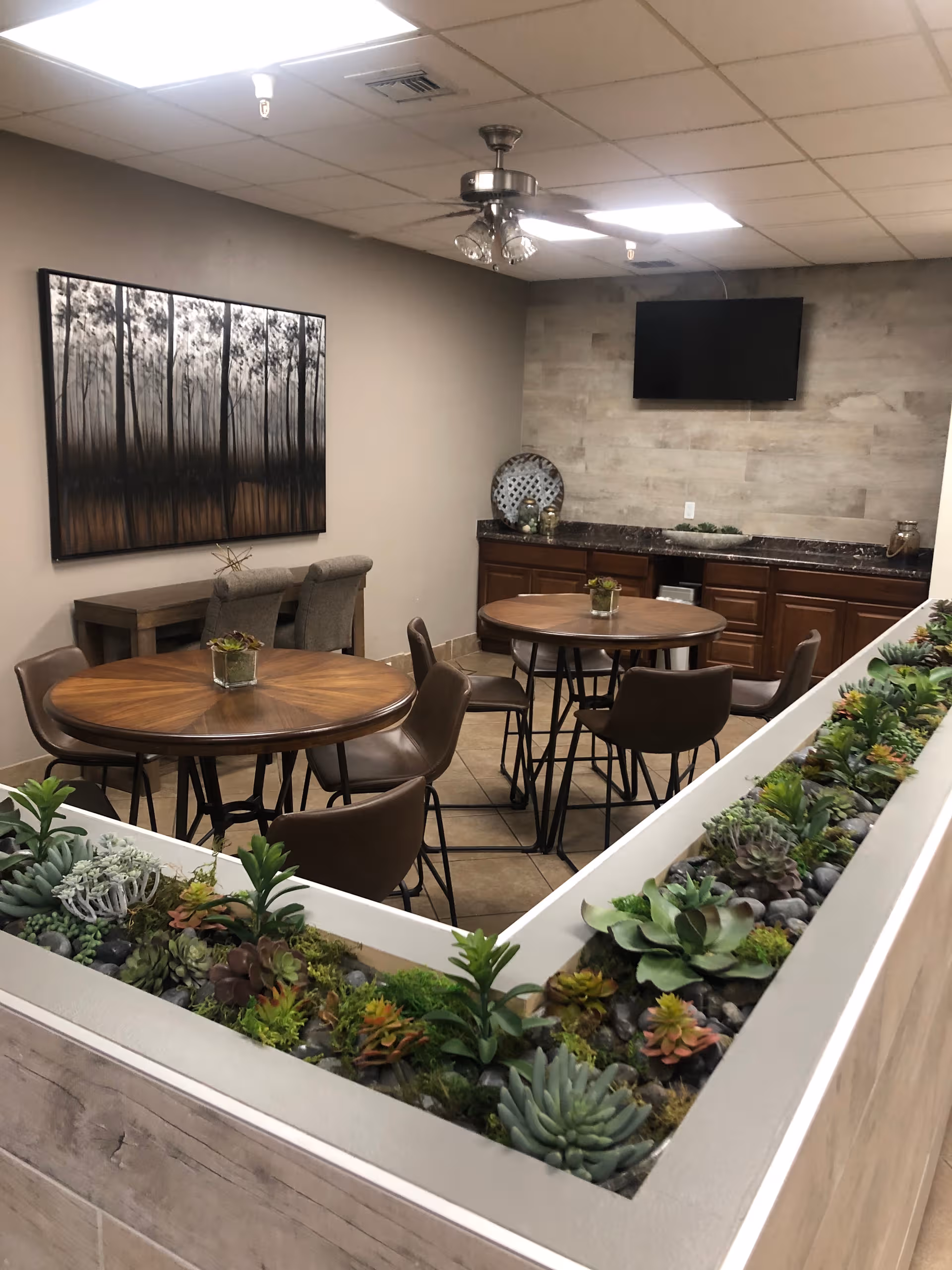 Indoor lounge with round tables, brown chairs, plant planter, and wall-mounted TV.