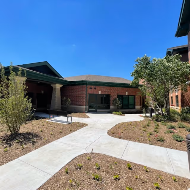Brick nursing facility entrance with a paved courtyard, walkways, benches, young landscaping, and a clear blue sky.