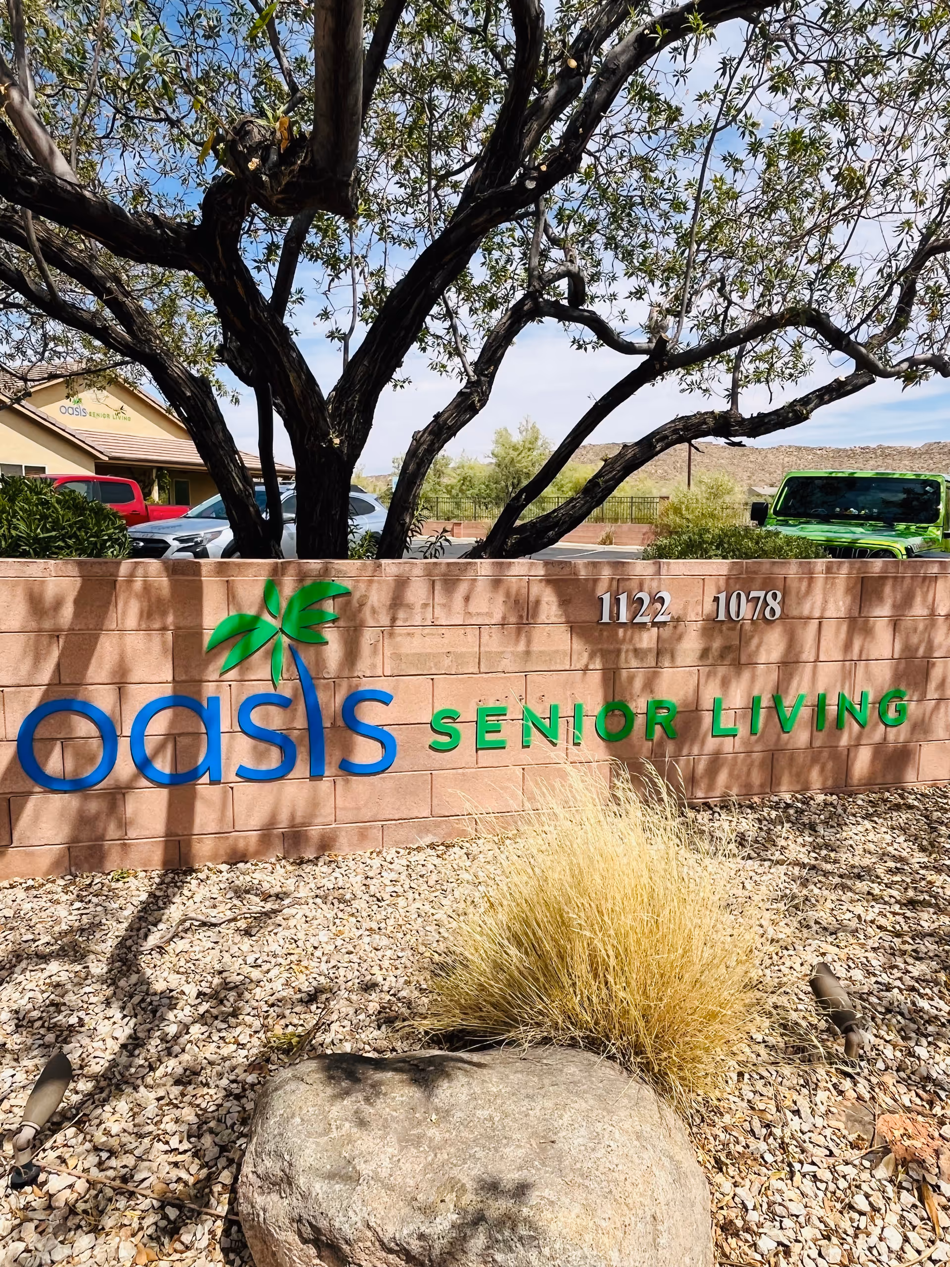 Front exterior sign reading Oasis Senior Living with tree, desert landscaping, and a parked vehicle.