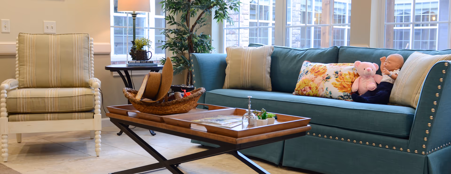 A cozy living room area with a teal sofa adorned with decorative pillows, including a floral one, and a baby doll and pink teddy bear sitting on it. Next to the sofa is a beige striped armchair. A wooden coffee table with a woven basket, a hat, and small decorative items sits in front of the seating. Large windows in the background let in natural light, and a tall green plant and a table lamp are also visible.