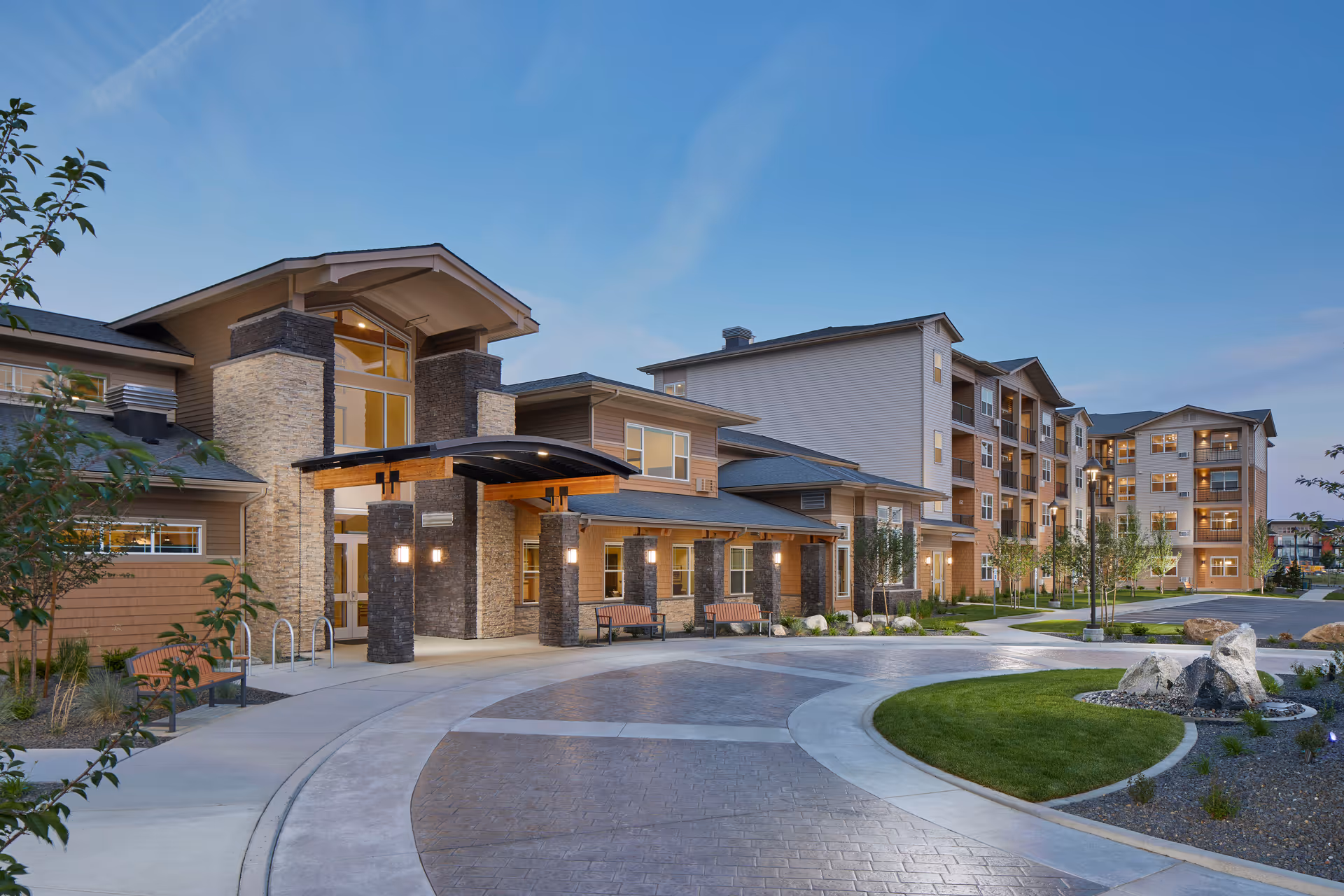 Exterior view of Revel Spokane senior living facility at dusk, showing a modern multi-story building with stone and wood accents, a covered entrance with benches, landscaped greenery, and a curved driveway.