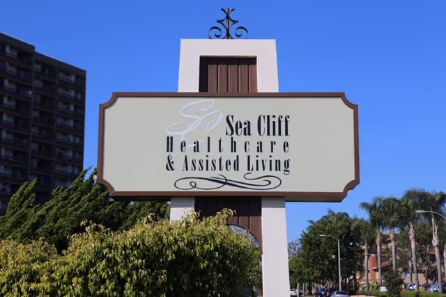 Outdoor sign for Sea Cliff Healthcare & Assisted Living with trees, a tall building, and a clear blue sky in the background.