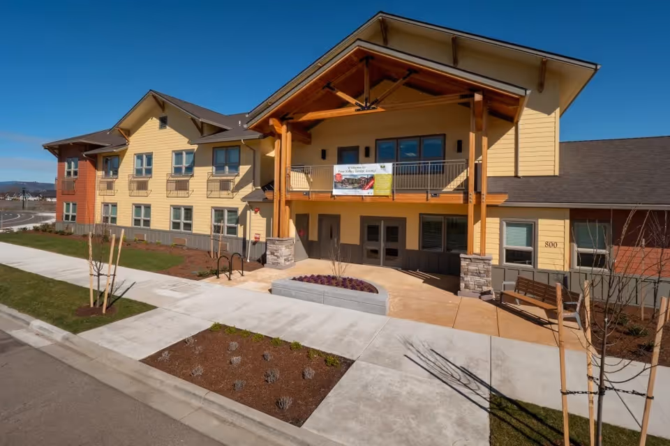 Front entrance of a two-story yellow and tan senior living building with a covered wooden porch, balcony, and landscaped walkway.