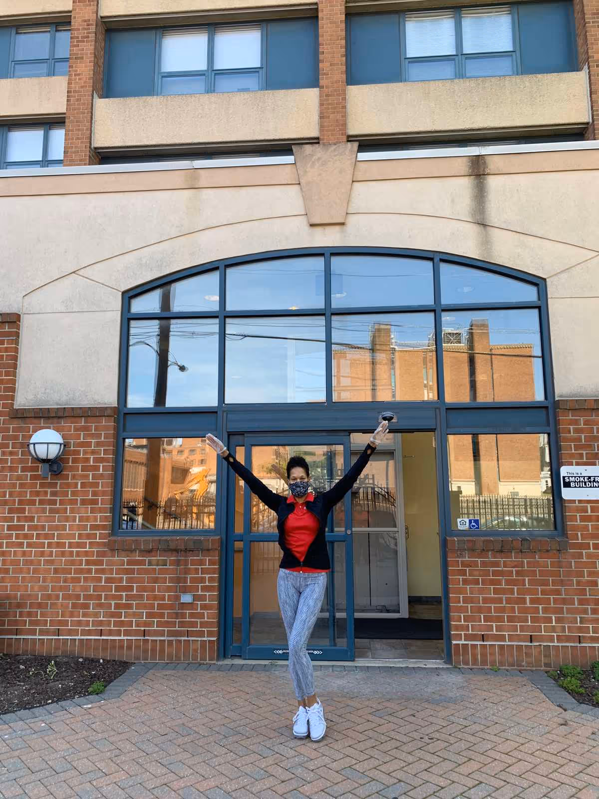 Person wearing a mask poses with arms raised at All American Home Care entrance.