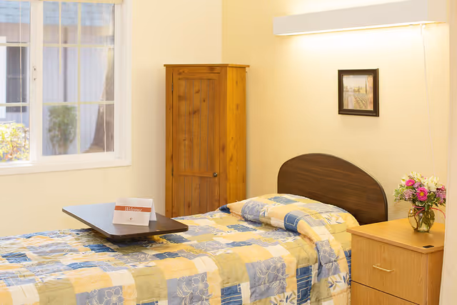 A single bed with a colorful patchwork quilt in shades of yellow, blue, and beige is placed against a light-colored wall. A wooden headboard is attached to the bed. Next to the bed is a wooden nightstand with a vase of fresh flowers and a small framed picture hanging above the bed. A wooden wardrobe stands in the corner near a window that lets in natural light. A small table with a welcome card is positioned on the bed.