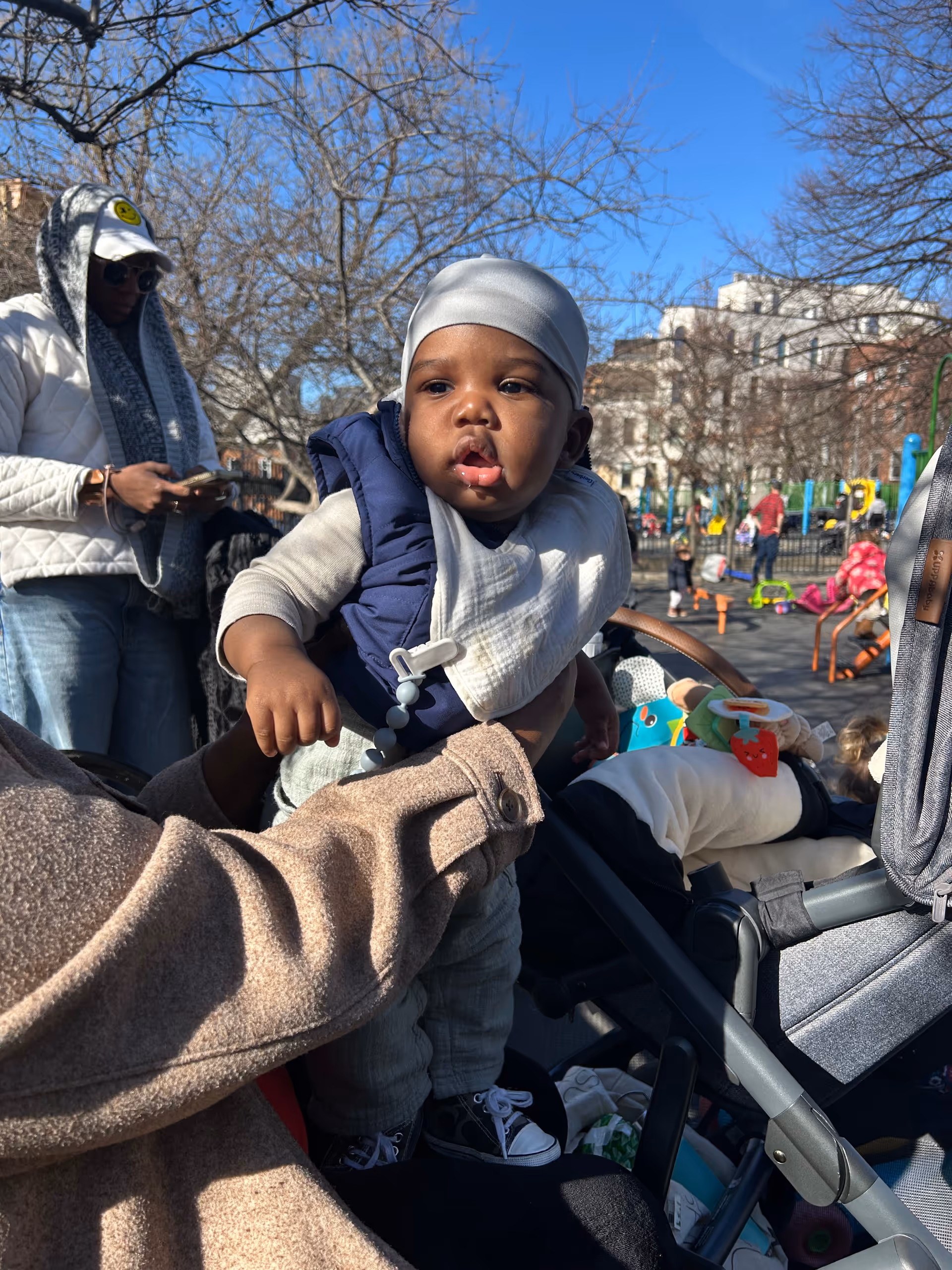 Baby in a stroller held by an adult outdoors at a park with playground equipment.