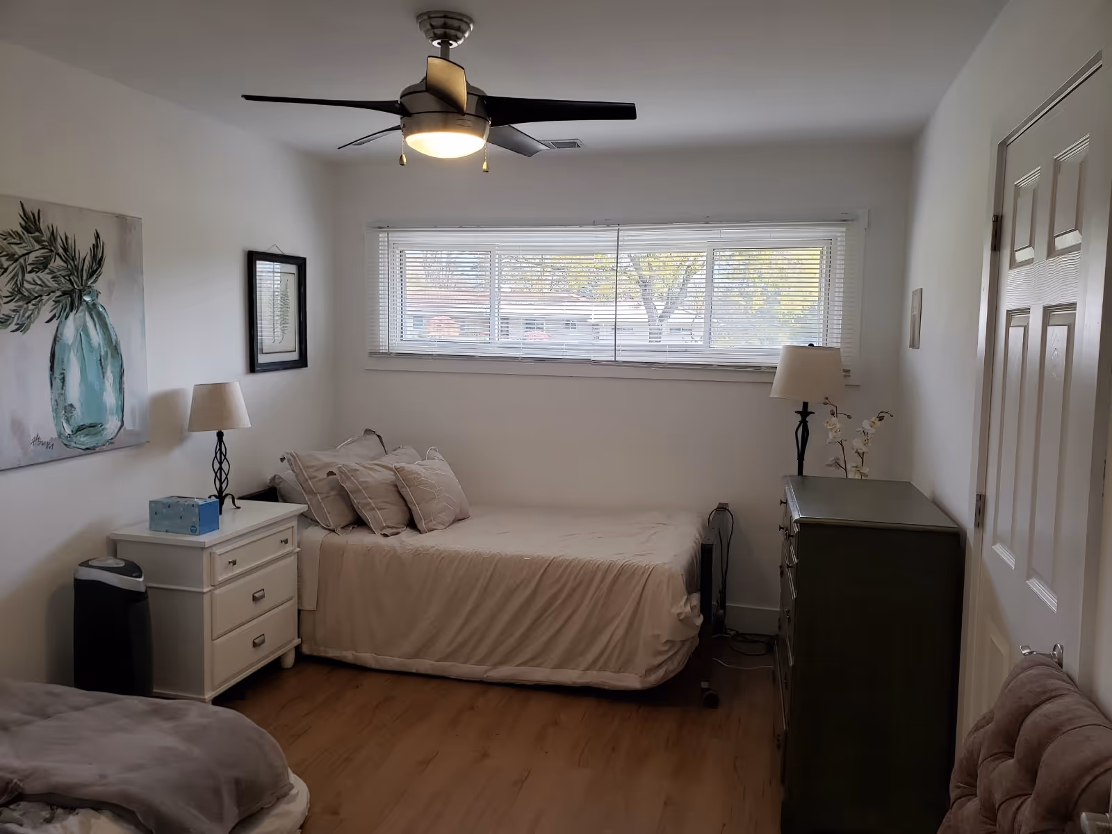 A tidy bedroom with two beds, nightstands, a dresser, and a large window with blinds.