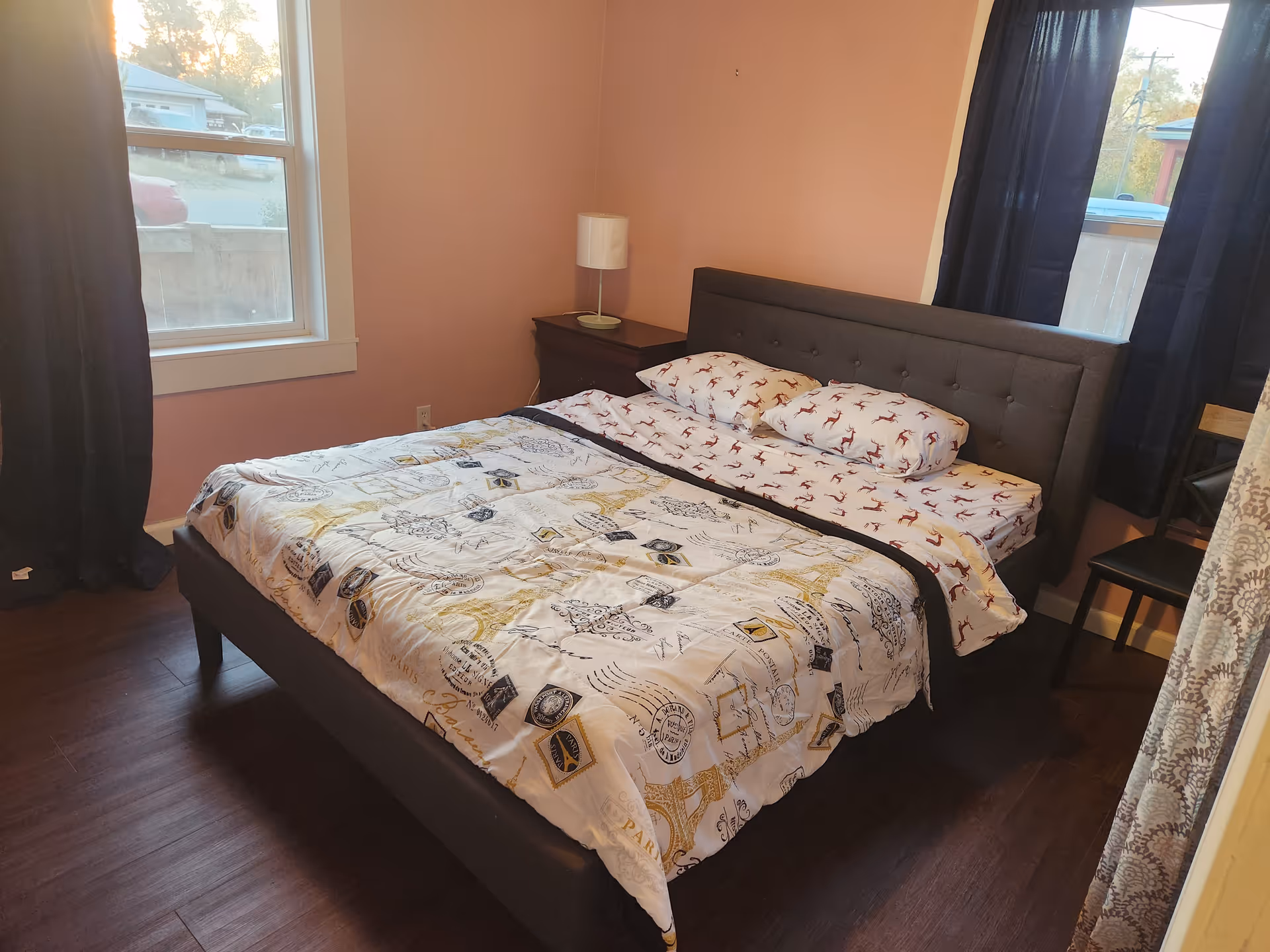 A furnished bedroom with a dark bed, nightstand, lamp, pink walls, and curtains.