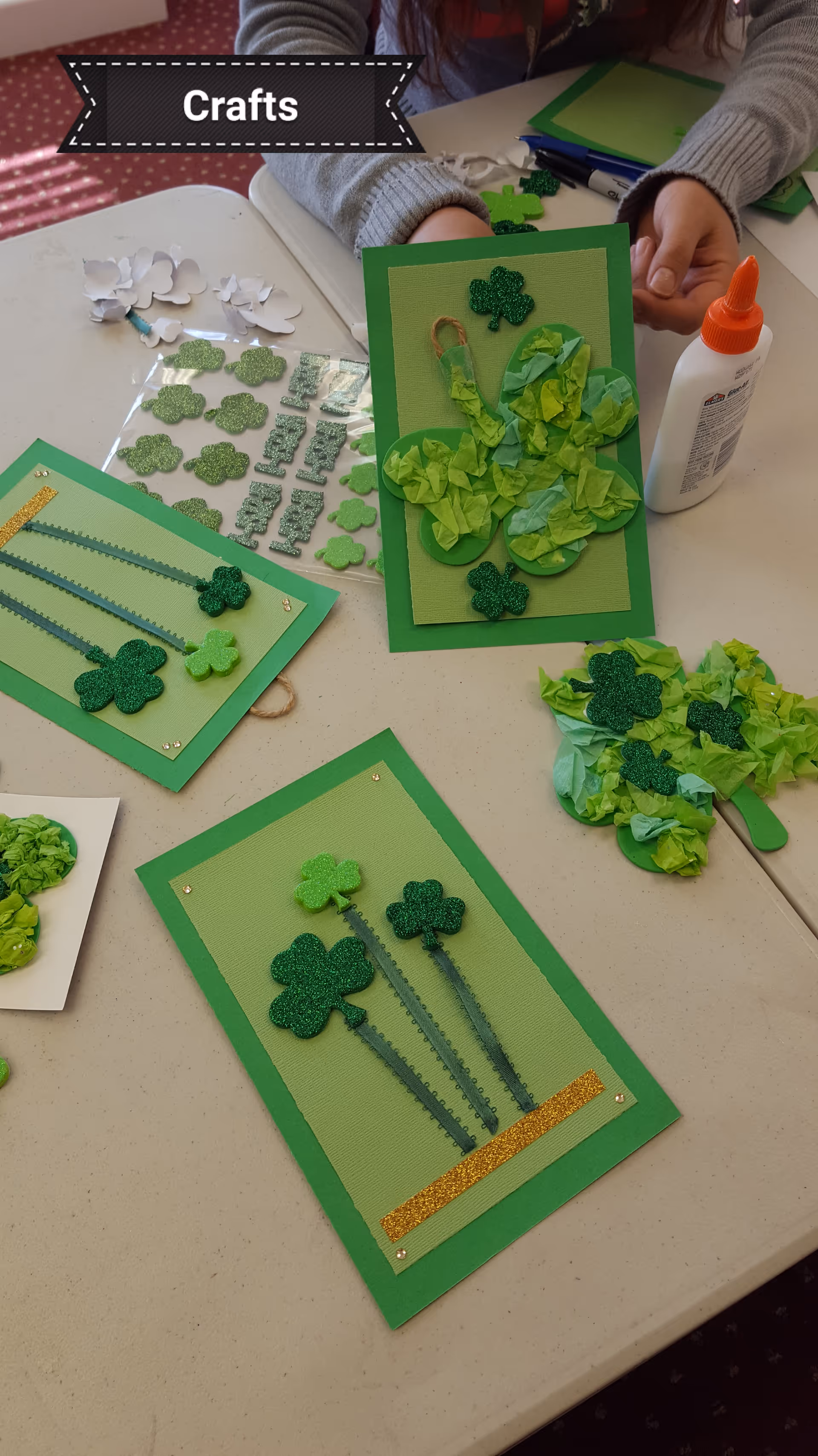 Indoor crafts table featuring green shamrock cards and visible hands crafting.