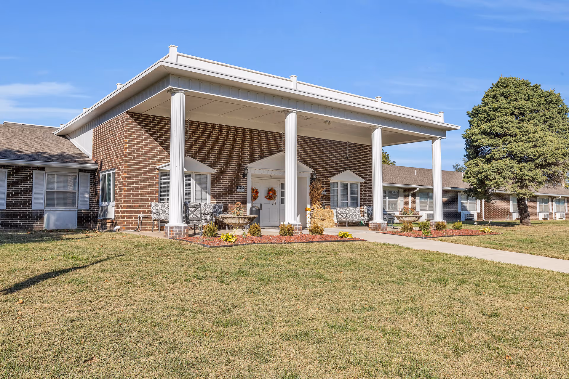 Exterior view of a single-story brick building with a covered entrance supported by four white columns. There are two benches with patterned cushions on either side of the entrance door, decorative plants, and a well-maintained lawn with a large tree to the right. The sky is clear and blue.