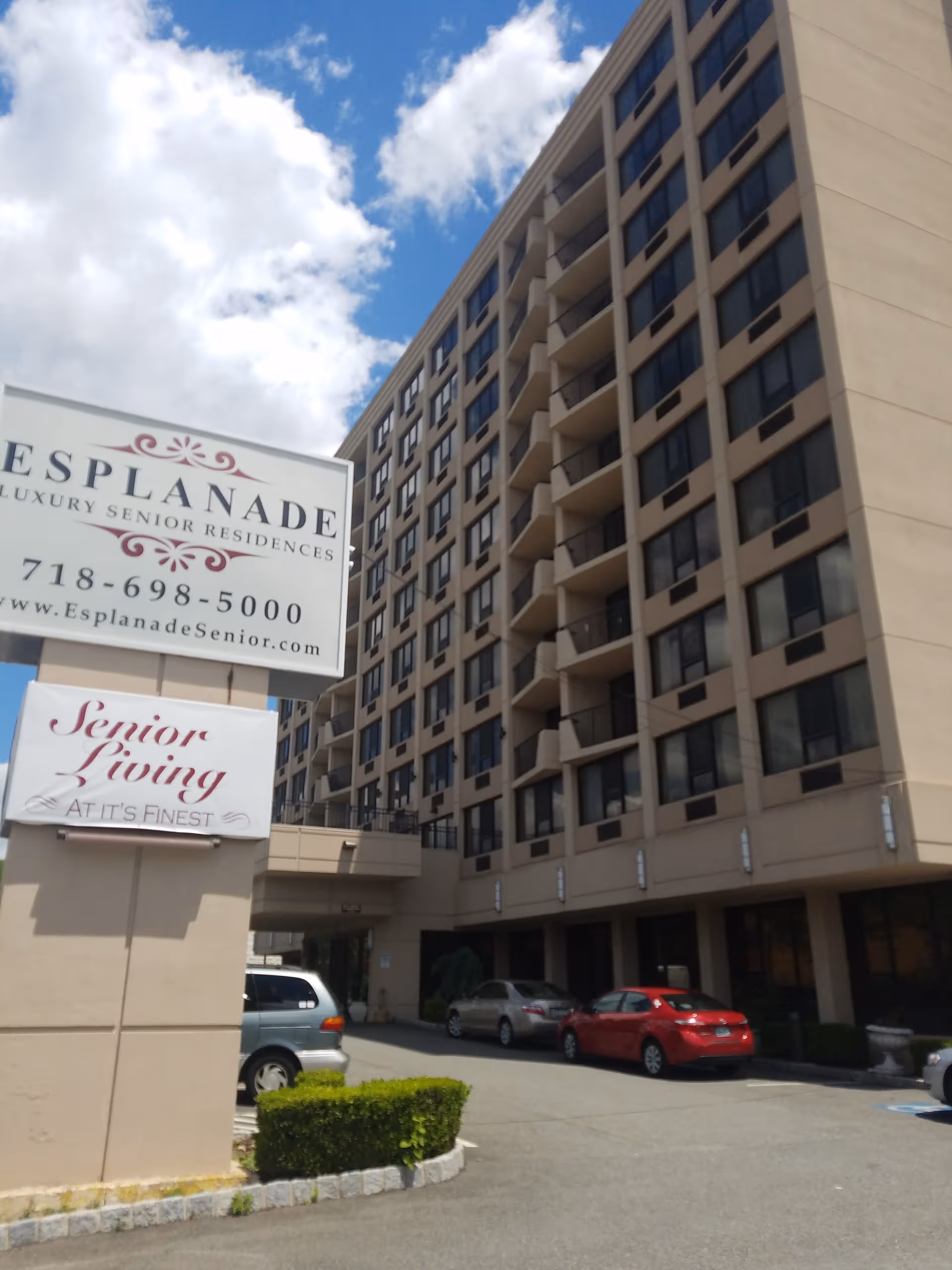 Tall beige senior residence building with signs, parked cars, and a clear blue sky.