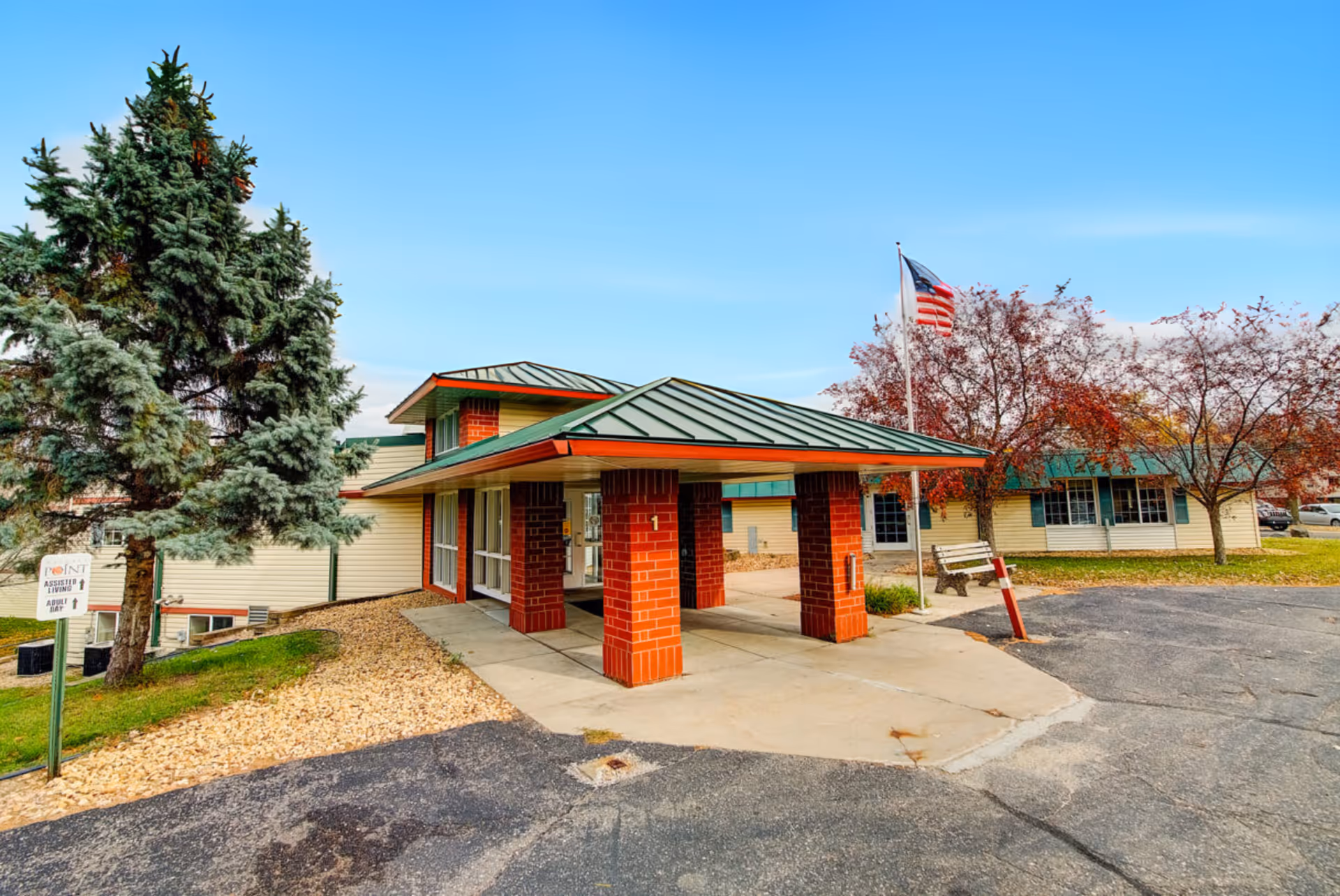Exterior view of Nature's Point Assisted Living facility showing a covered entrance with red brick pillars and a green metal roof. There are trees with autumn foliage, an American flag on a flagpole, a bench, and a paved driveway in front of the building under a clear blue sky.