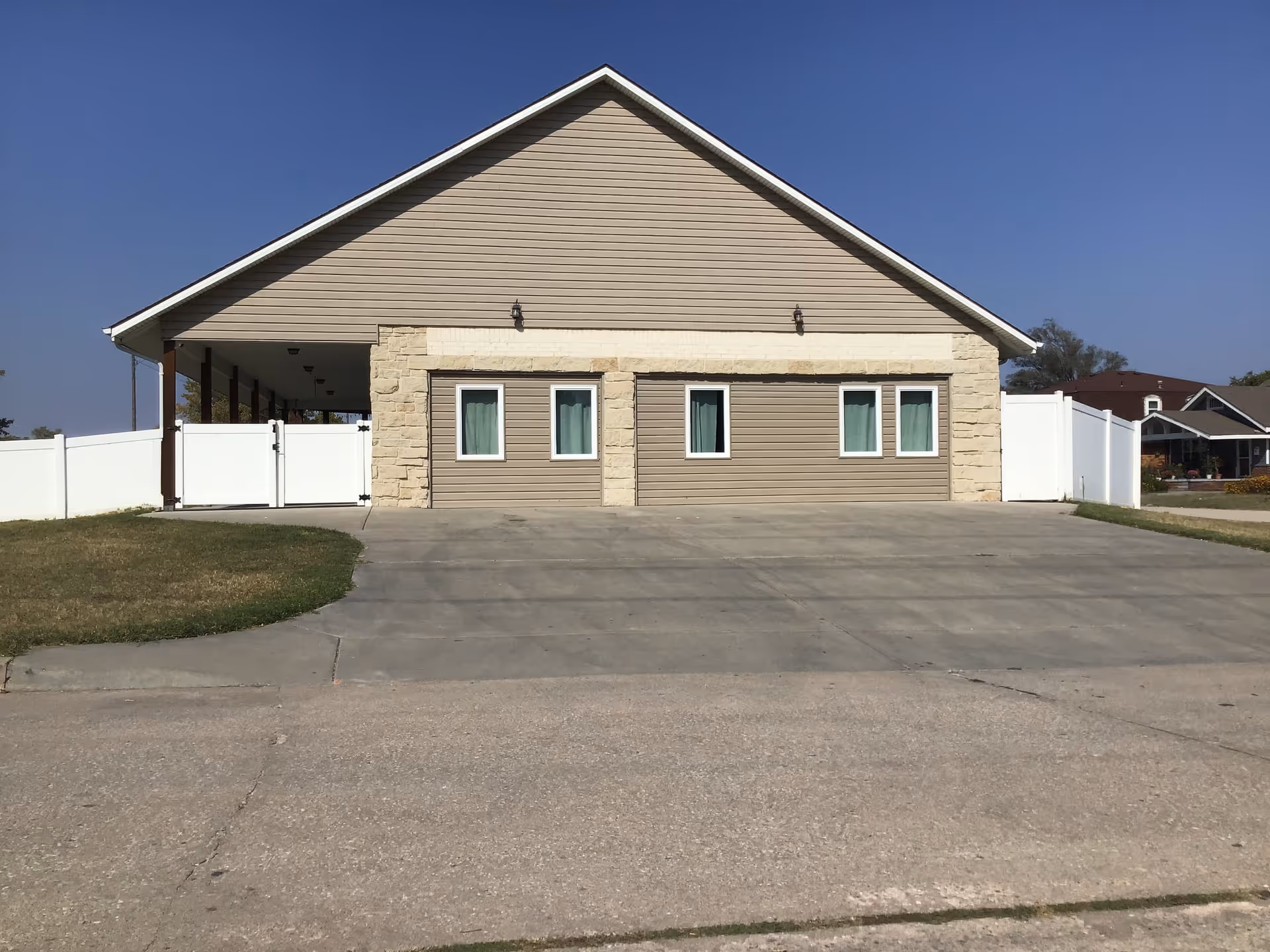 Front exterior view of a single-story building with beige siding and stone accents, four small windows with curtains, a large concrete driveway, and white fencing on both sides under a clear blue sky.