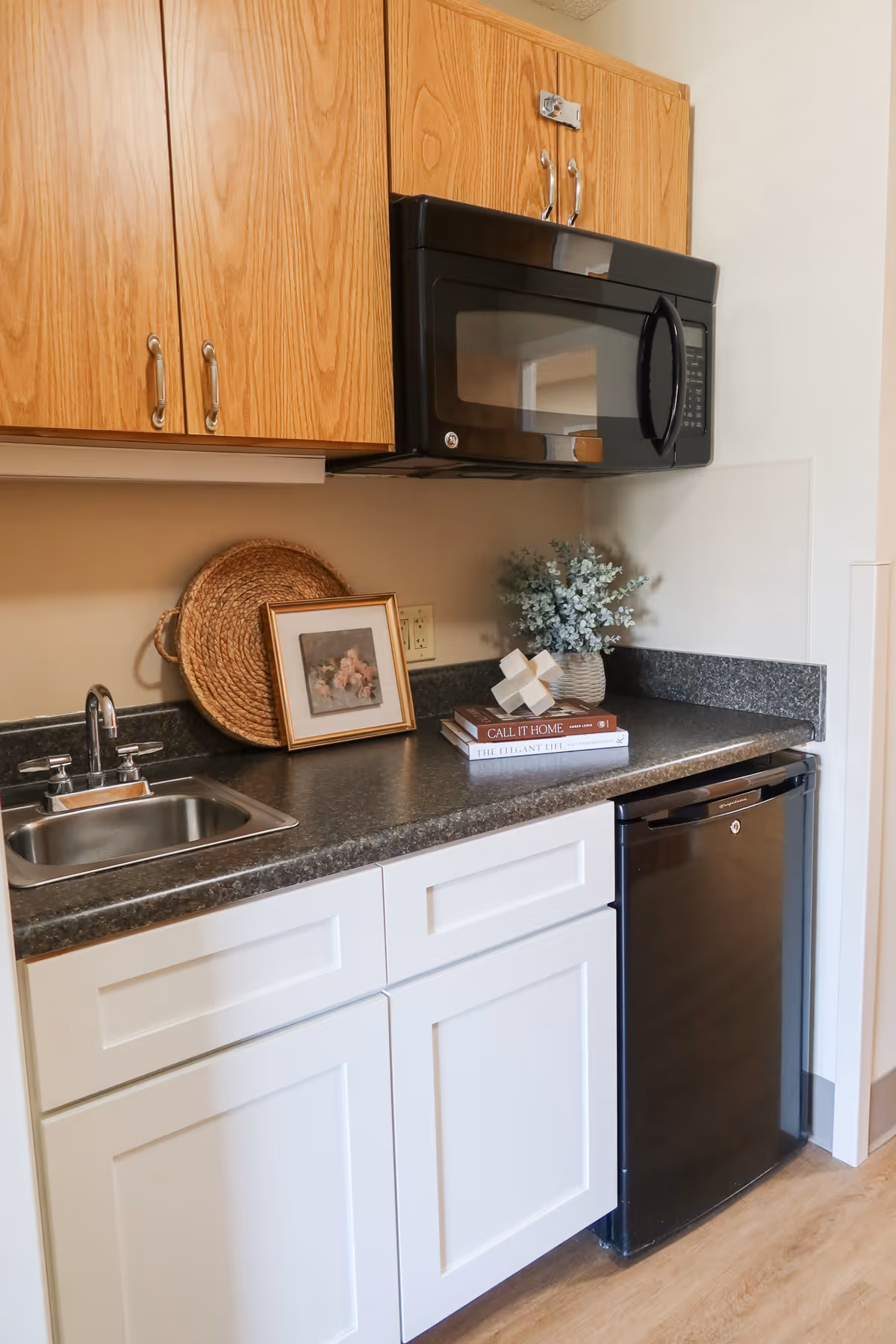 Compact kitchen with wooden cabinets, microwave, sink, and decorative items on counter.