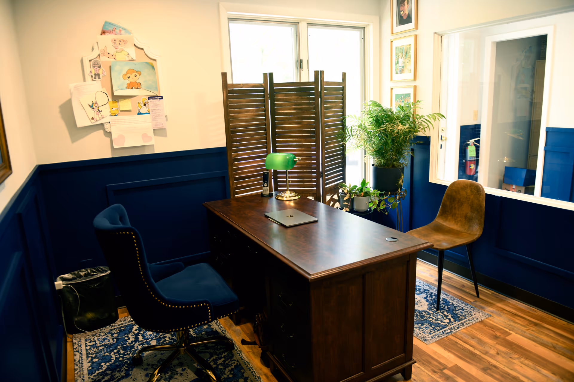 A wood desk with chairs, plants, and partition in a small office inside a senior facility.