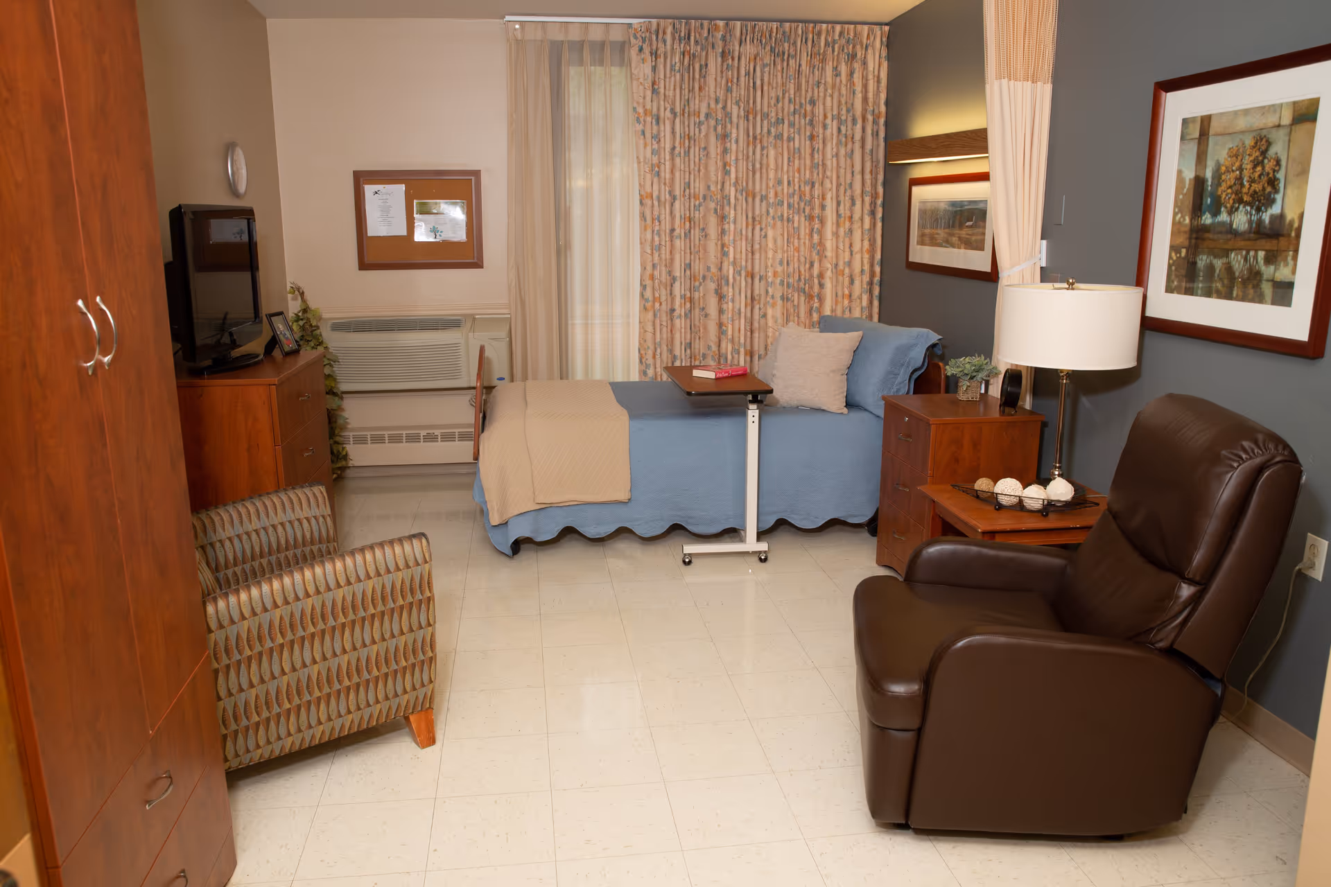 A cozy room with a single bed covered in a blue blanket and beige throw, a small overbed table with a book on it, a wooden nightstand with a lamp and decorative items, a brown leather recliner chair, a patterned upholstered chair, a wooden dresser with a TV on top, and framed artwork on the walls. The window is covered with floral curtains.