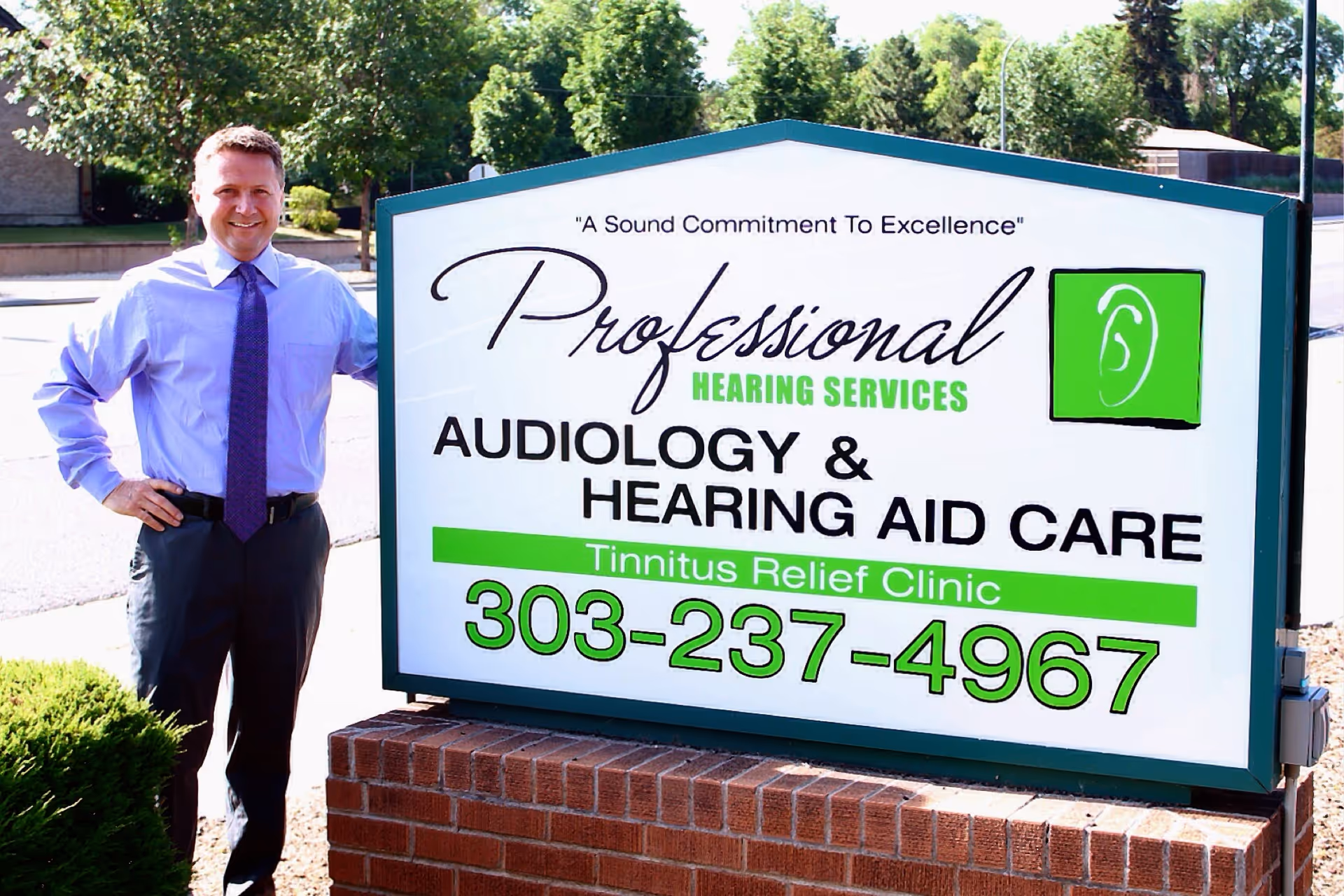 Man stands beside a large outdoor sign for Professional Hearing Services.