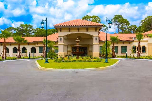 Front exterior view of Discovery Commons South Biscayne facility featuring a circular driveway with a central landscaped island and a tiered water fountain. The building has a beige facade with arched windows and a red tile roof, surrounded by palm trees and greenery under a partly cloudy blue sky.