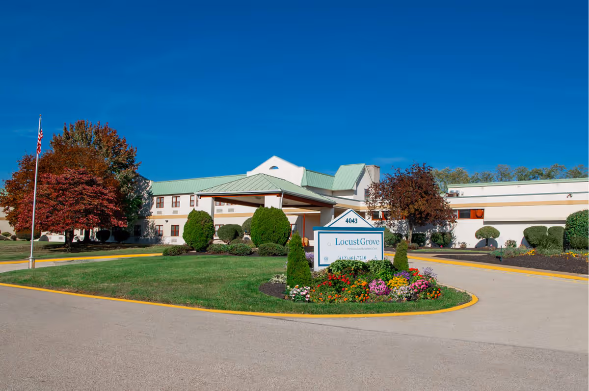 Exterior view of Locust Grove Personal Care & Memory Care facility with a driveway, landscaped garden with colorful flowers, trimmed bushes, and trees with autumn foliage under a clear blue sky.