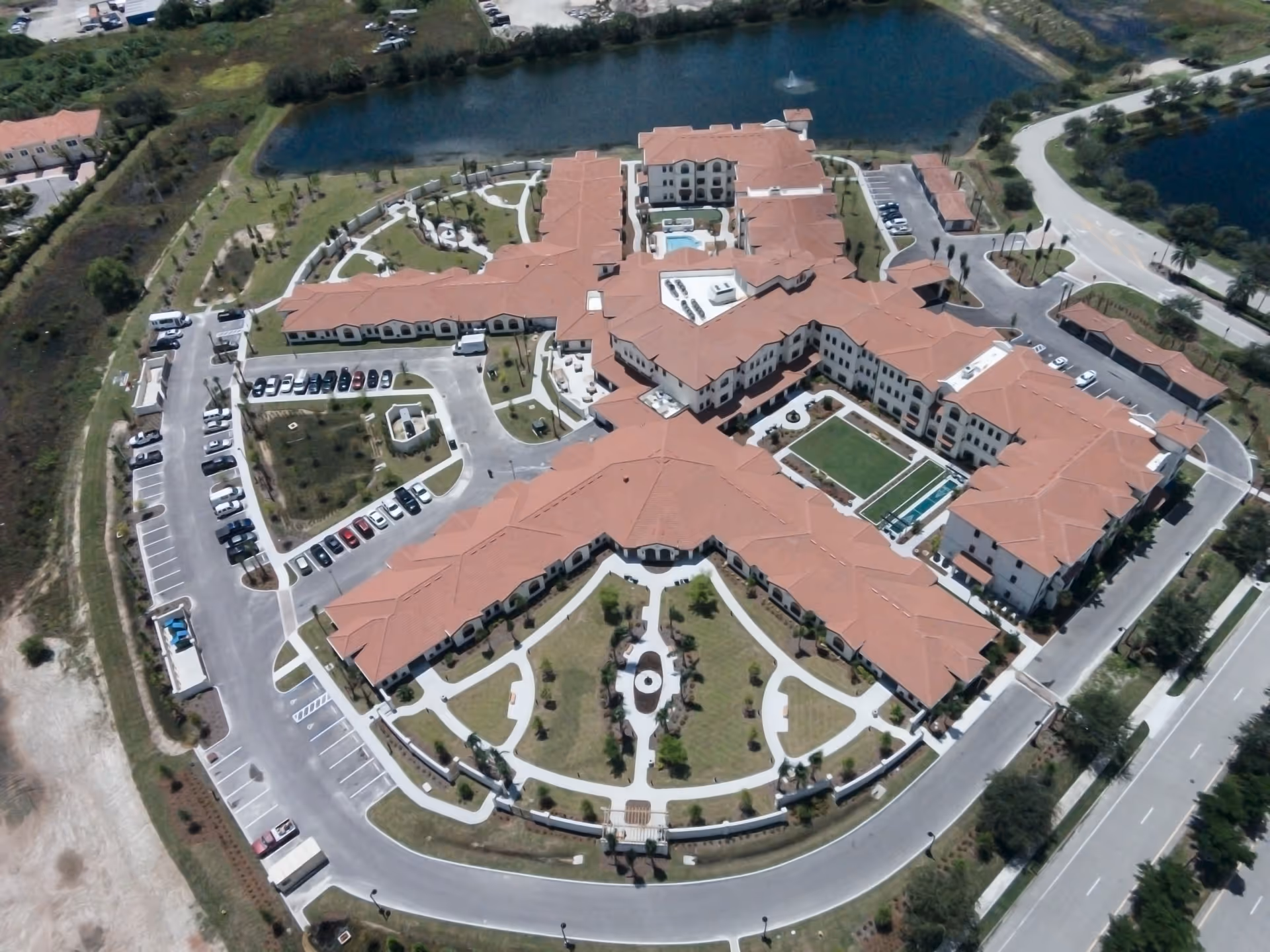 Aerial view of the American House Coconut Point senior living facility showing a large building complex with red-tiled roofs, surrounding landscaped gardens, parking lots, and a pond with a fountain in the background.