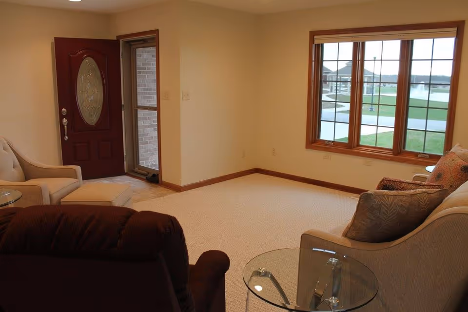 Cozy living room with armchairs, a glass side table, a red front door, and a large window overlooking a courtyard and fountain.