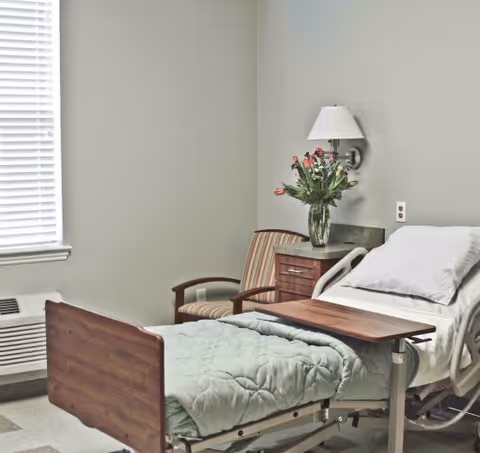 Hospital-style patient bed in a quiet bedroom with flowers, lamp, and chair.
