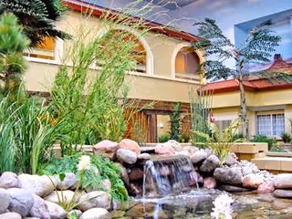 Outdoor courtyard area at Viewcrest Health Center featuring a small waterfall flowing over rocks into a pond surrounded by plants and greenery, with the building's beige exterior and arched windows in the background.