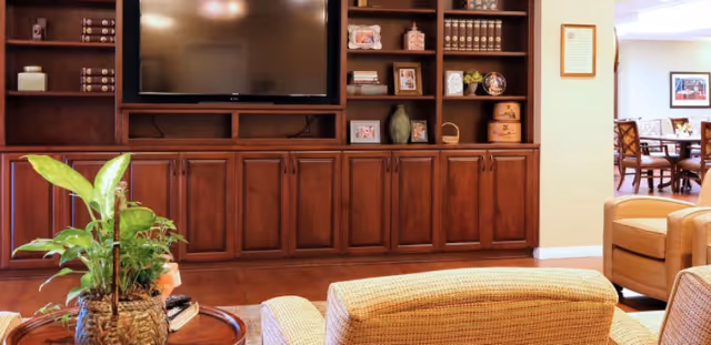 Cozy common room with a large wooden built-in entertainment center and TV, upholstered chairs, a coffee table with a potted plant, and a view into a dining area.