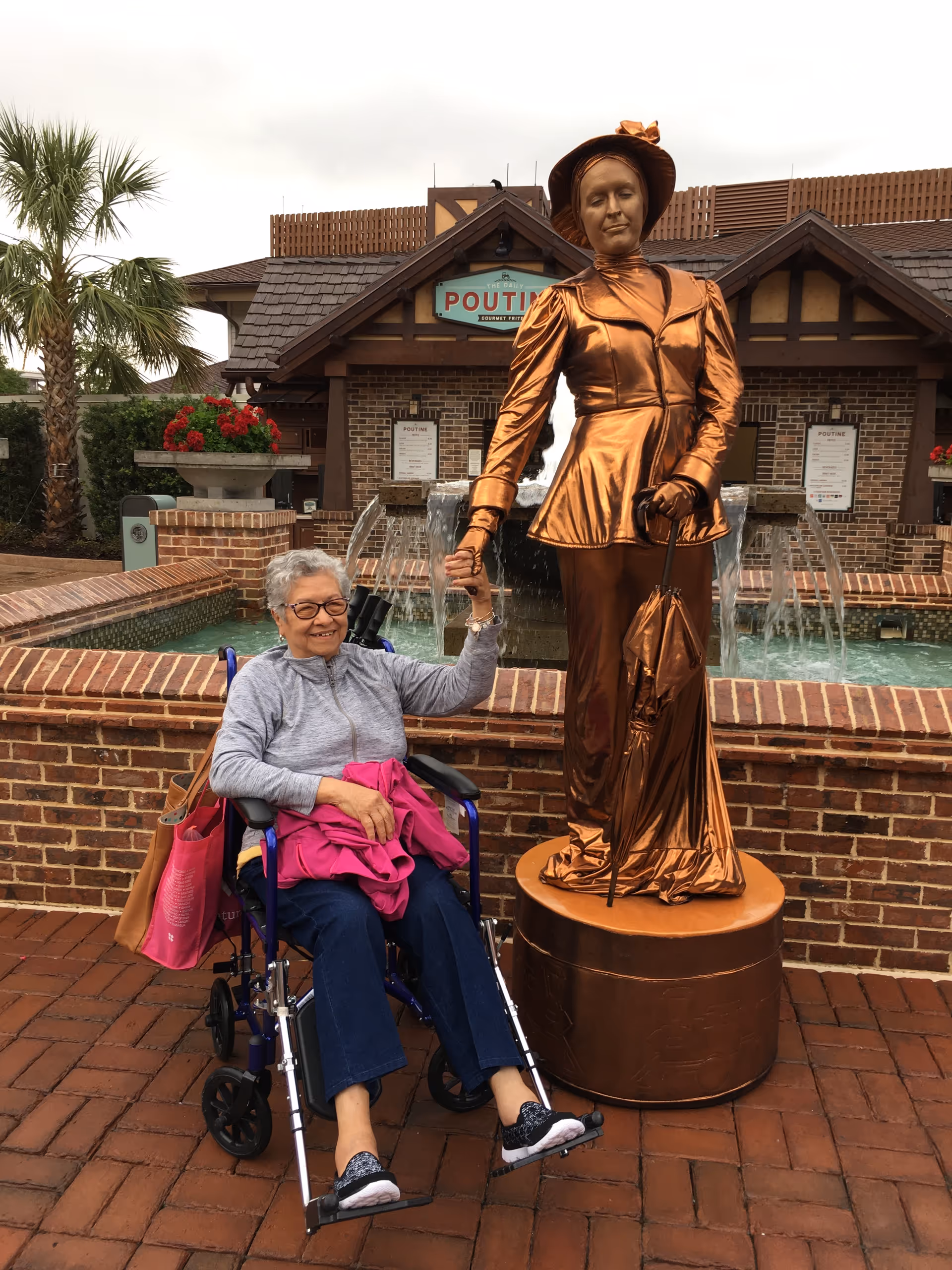 Elderly woman in a wheelchair grasps the bronze statue's hand outdoors near a fountain.