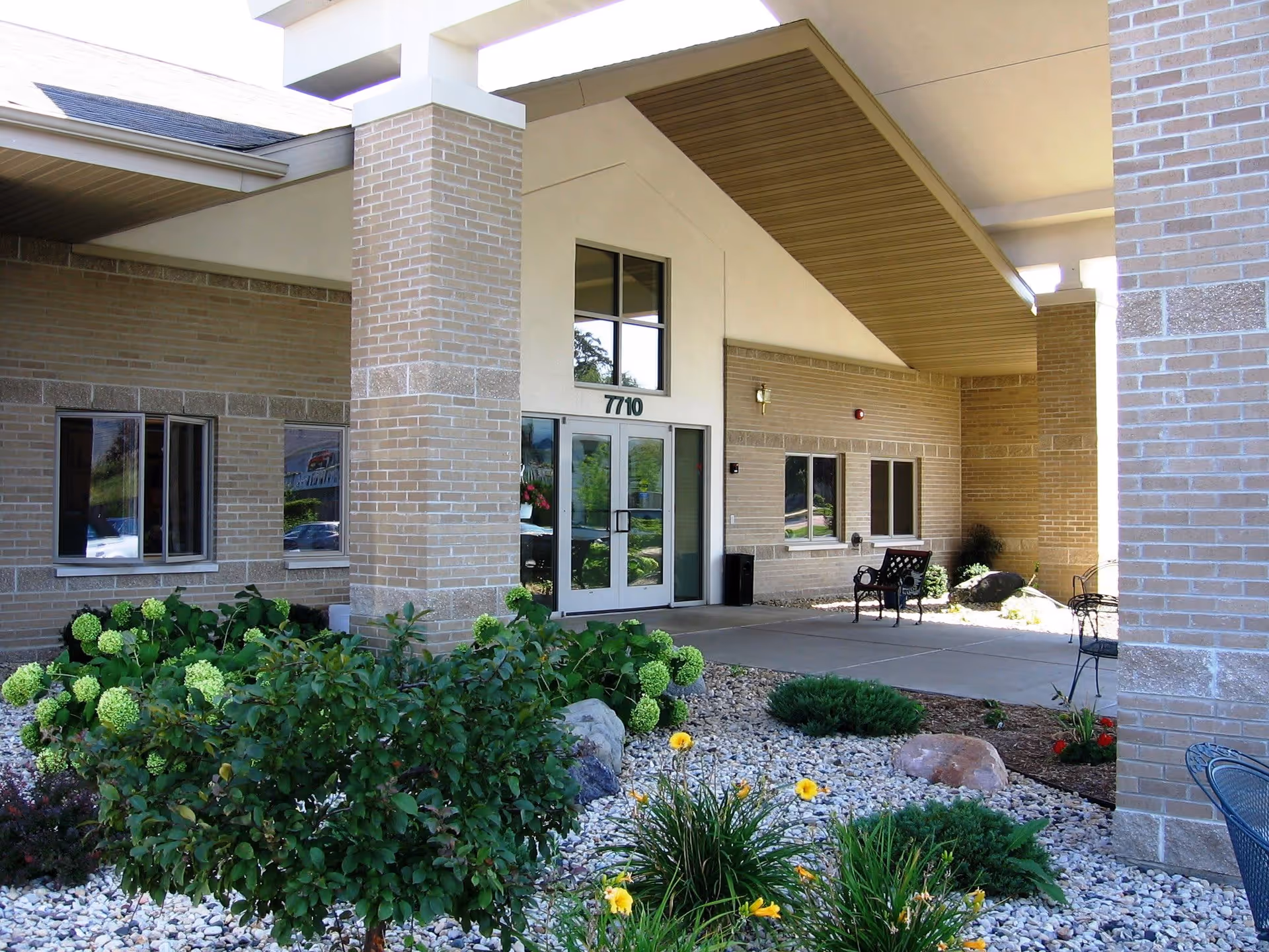Entrance of a building with beige brick walls and a covered walkway supported by large pillars. There are glass double doors with the number 7710 above them. In front of the entrance, there is a landscaped area with green bushes, flowering plants, and rocks. A bench and a chair are placed near the wall under the covered area.
