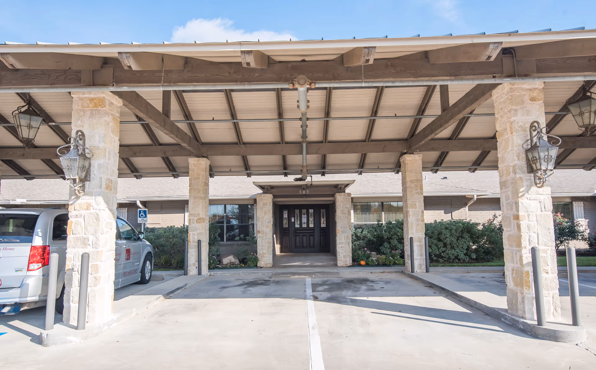 Covered entrance area of a senior living facility with stone pillars and a dark wooden door in the background. There is a parked van on the left side and some greenery along the building facade.
